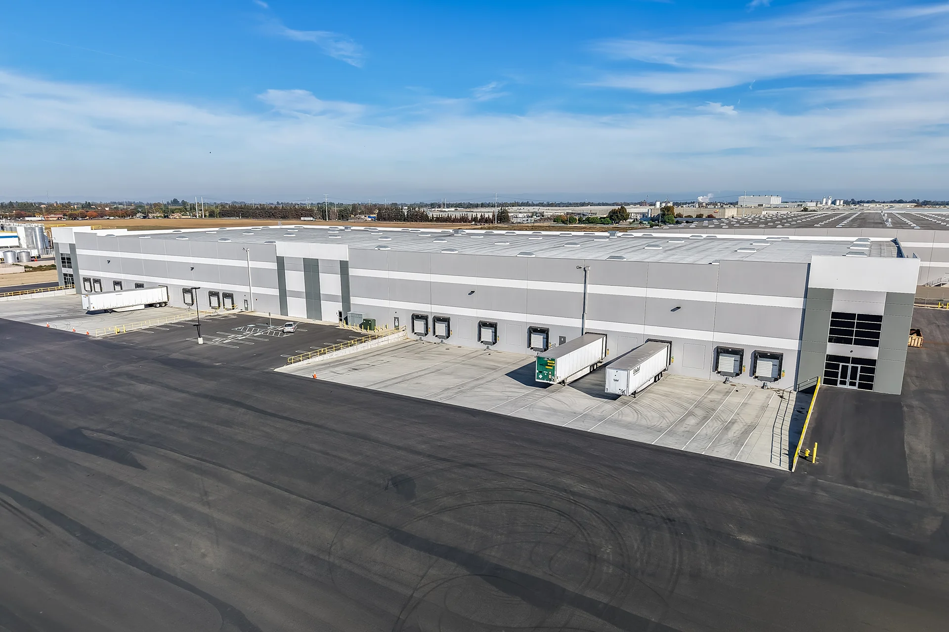 Panoramic drone view tracking along the side of an institutional industrial complex, detailing alternating light and dark grey elastomeric wall coatings, specialized dock canopy lighting, grade-level drive-in doors, and regional distribution infrastructure visible in the background.