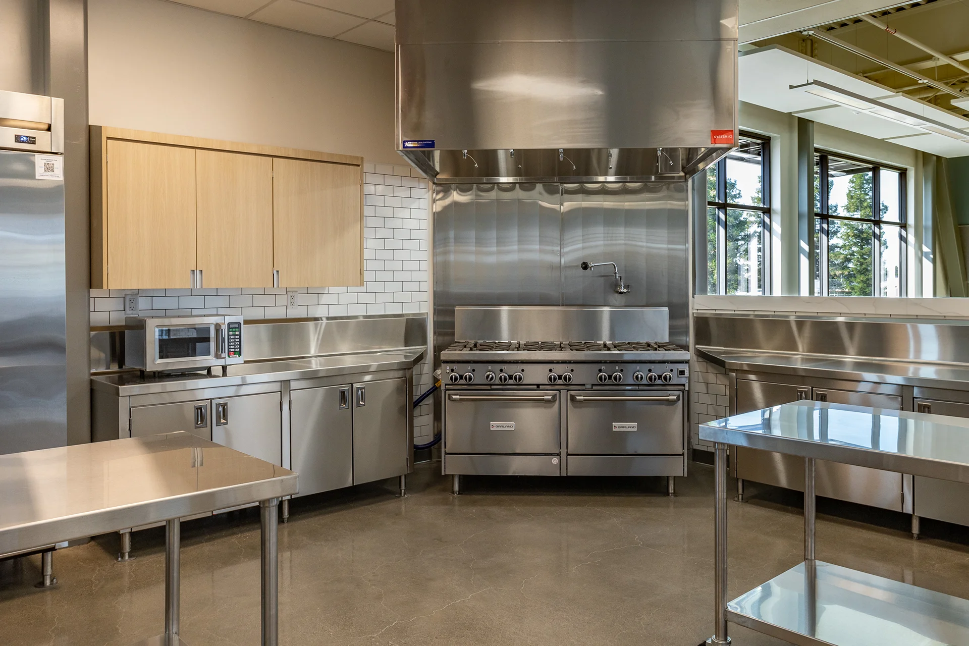 Wide view of a modern high school culinary lab hot line featuring an integrated microwave nook, stainless steel base units, gas range equipment, and open sightlines to large perimeter windows.