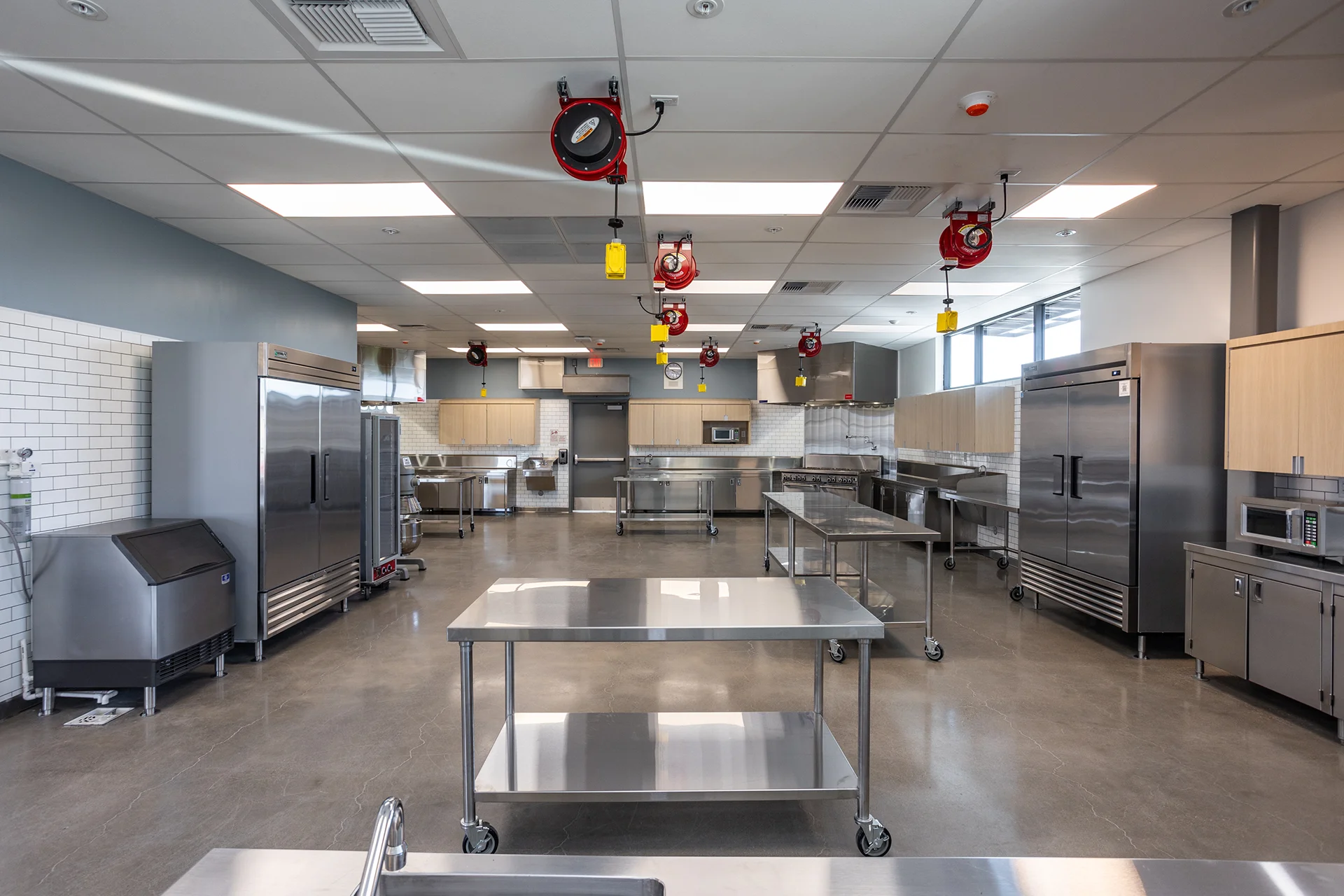 Symmetry shot down a commercial-grade culinary lab showcasing parallel rows of stainless steel prep tables on wheels, double-door reach-in refrigerators, an industrial ice machine, and ceiling-suspended spring-loaded retractable power cords.