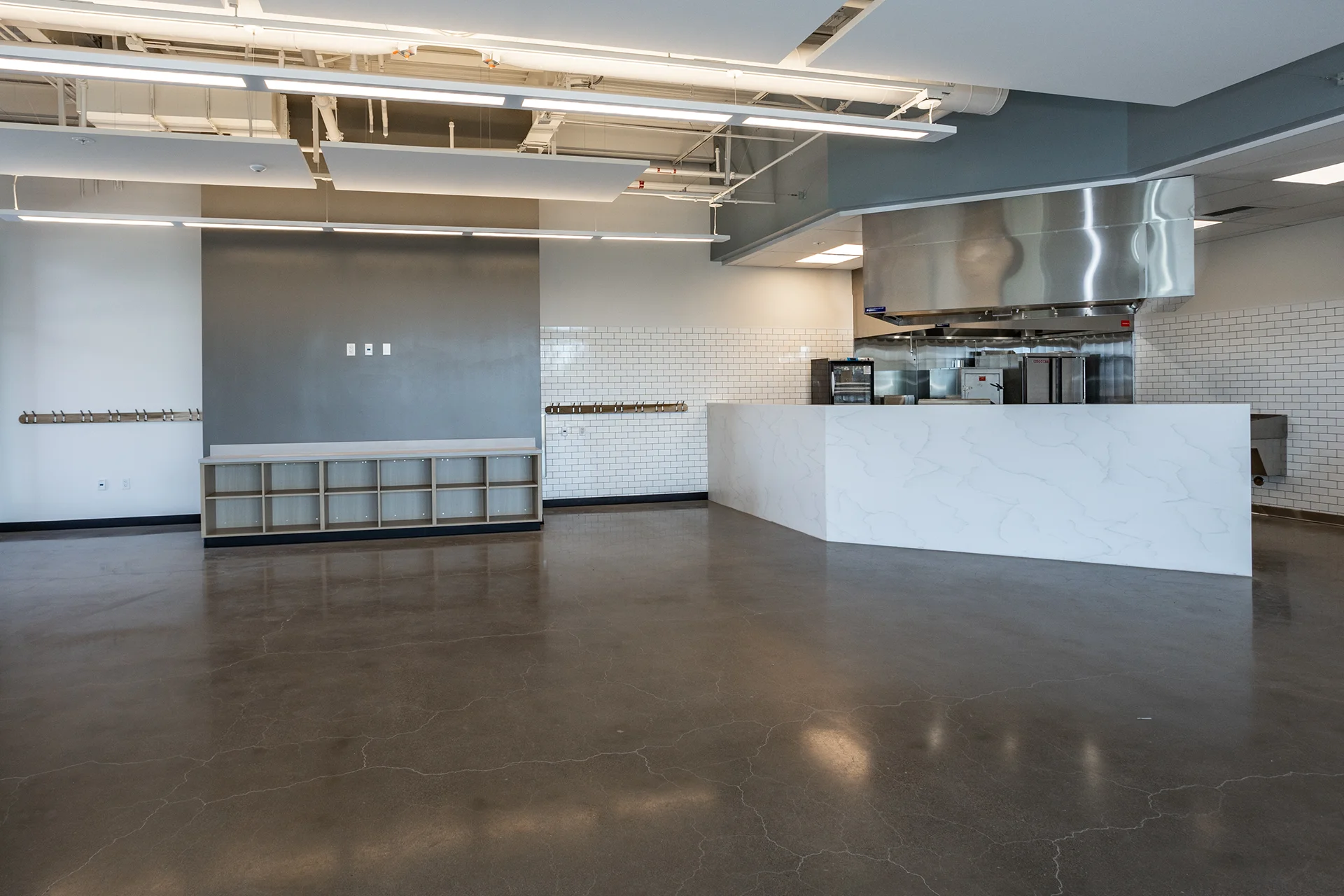 Wide-angle view of a modern culinary training center showing a large white faux-marble waterfall service counter, white subway-tiled partition walls, custom open millwork, and exposed overhead utility piping.