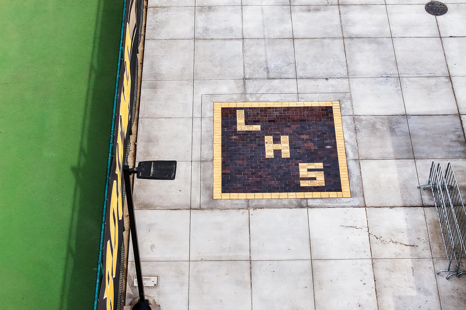 Top-down view of a custom dark brick paver inlay spelling LHS on a concrete courtyard next to a tennis court.