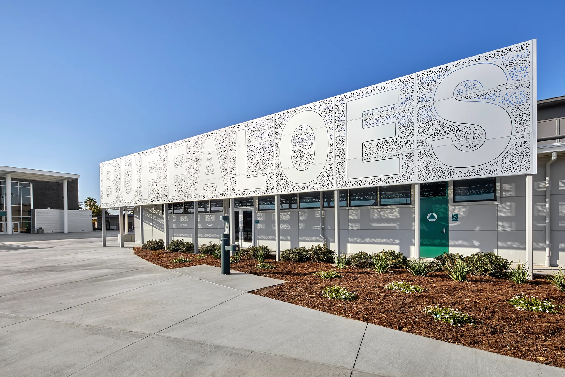 Wide angle exterior shot of a campus building in Manteca, California, featuring an expansive white laser-cut perforated metal sunscreen panel spelling out a mascot name, set over dark green trim accents, concrete flatwork, and organic bark mulch landscaping.