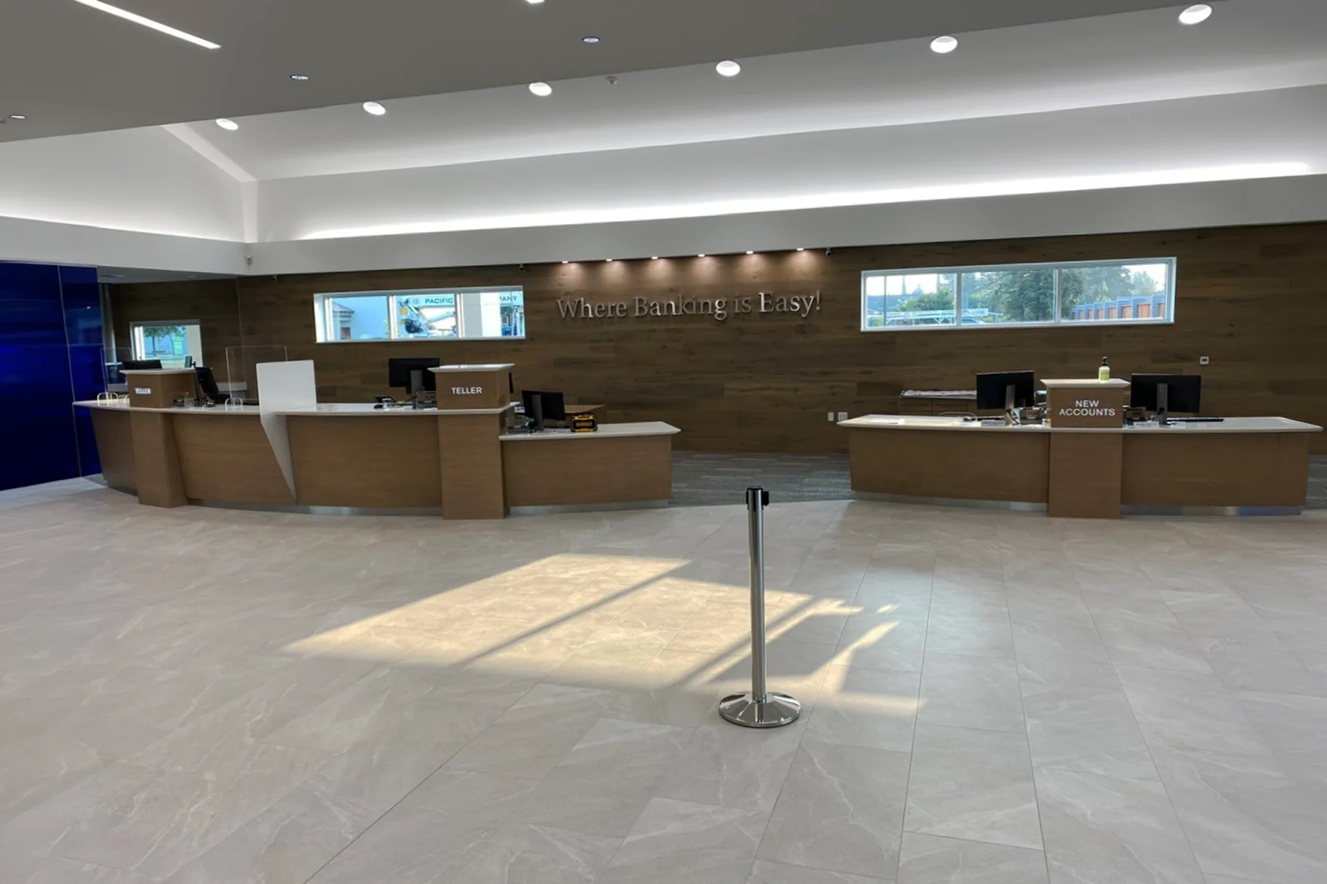Main customer service area inside the retail bank, featuring light oak teller pods, white solid-surface countertops, large format stone tile floors, and a horizontal wood-paneled accent wall displaying the metallic silver phrase Where Banking is Easy.