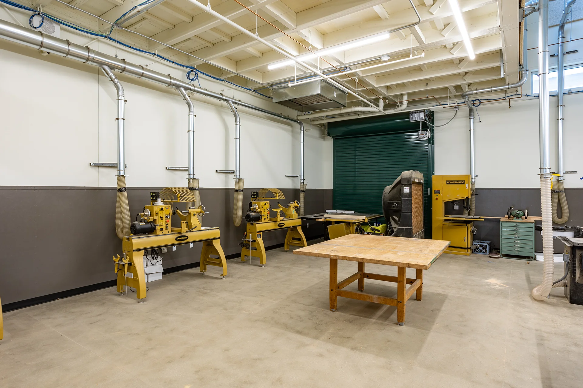 Specialized vocational training room interior featuring two yellow Powermatic wood lathes with clear safety hoods, a central maple layout table, a commercial metal rolling door, and custom flexible source extraction ventilation systems secured to plaster walls.