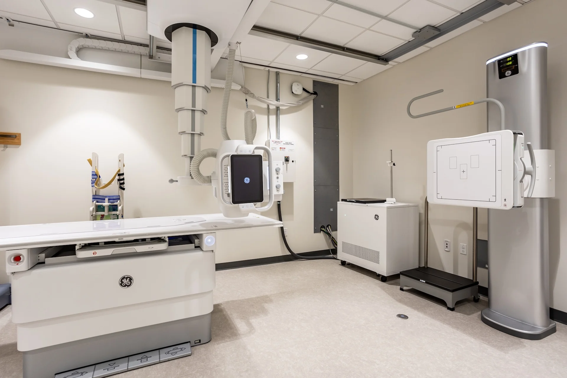 Advanced medical imaging room showcasing a GE digital radiography machine, an adjustable ceiling-mounted X-ray tube suspension arm, an elevate-capable examination table, a vertical wall bucky detector stand, and shielded power systems.