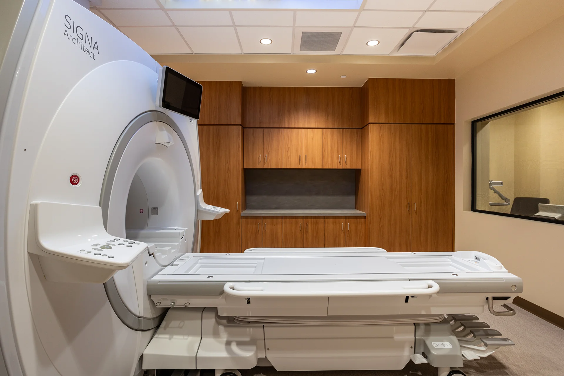 Interior view of a medical imaging suite featuring a GE SIGNA Architect MRI scanner and patient table, integrated wood-grain storage cabinetry with a solid-surface counter, and a lead-lined control room observation window beneath recessed acoustic ceiling tiles.