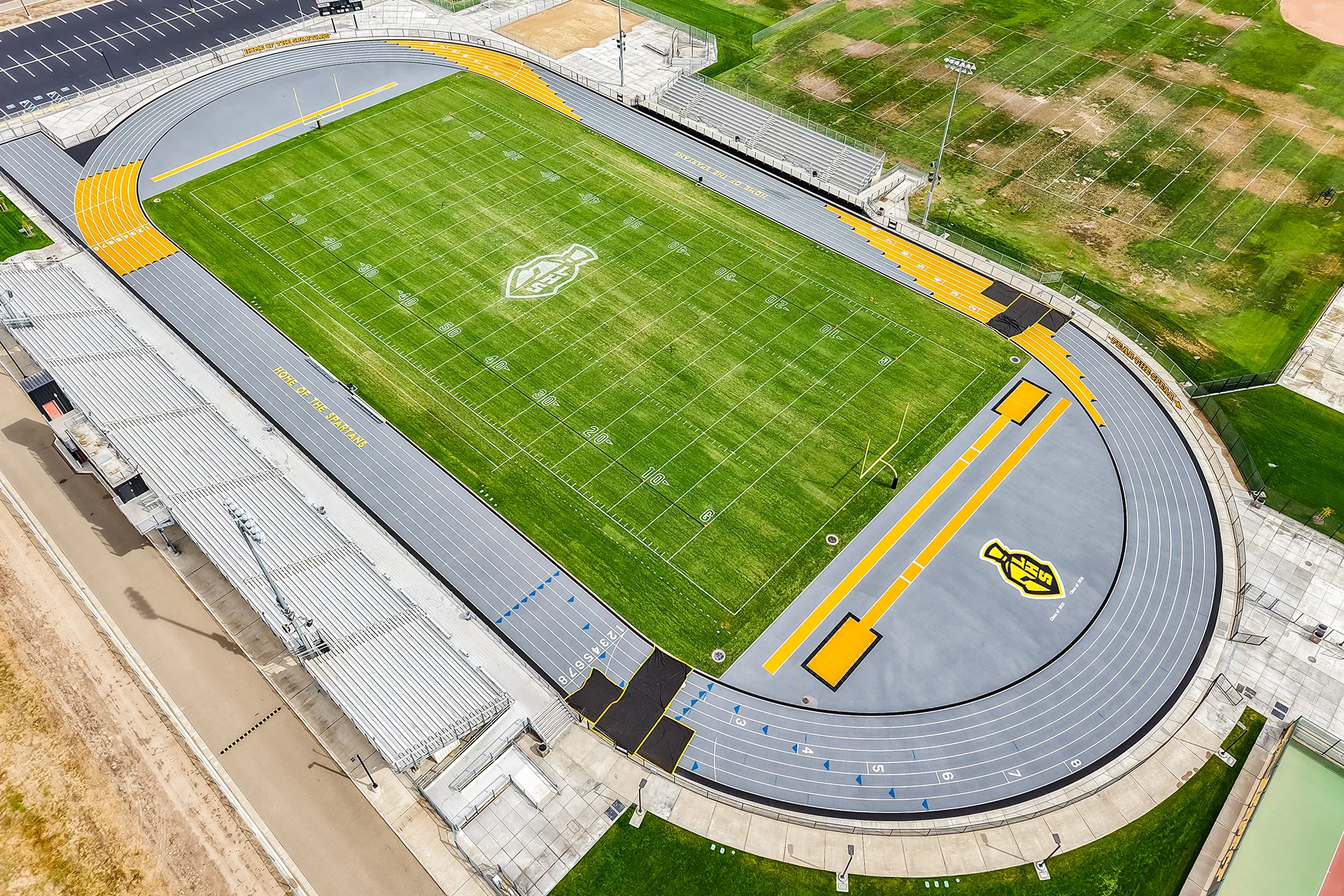High-angle diagonal drone view showing the complete football stadium layout, aluminum bleachers, and a custom grey running track.