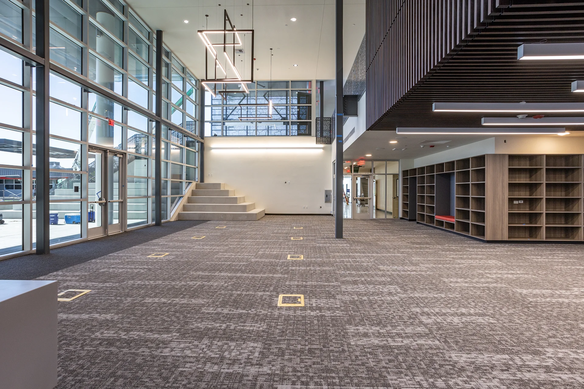 Large campus common area in Manteca, California boasting a floor-to-ceiling aluminum storefront glass curtain wall, industrial steel framing, a wood-slat acoustic ceiling accent, suspended custom LED chandeliers, and built-in millwork lockers.