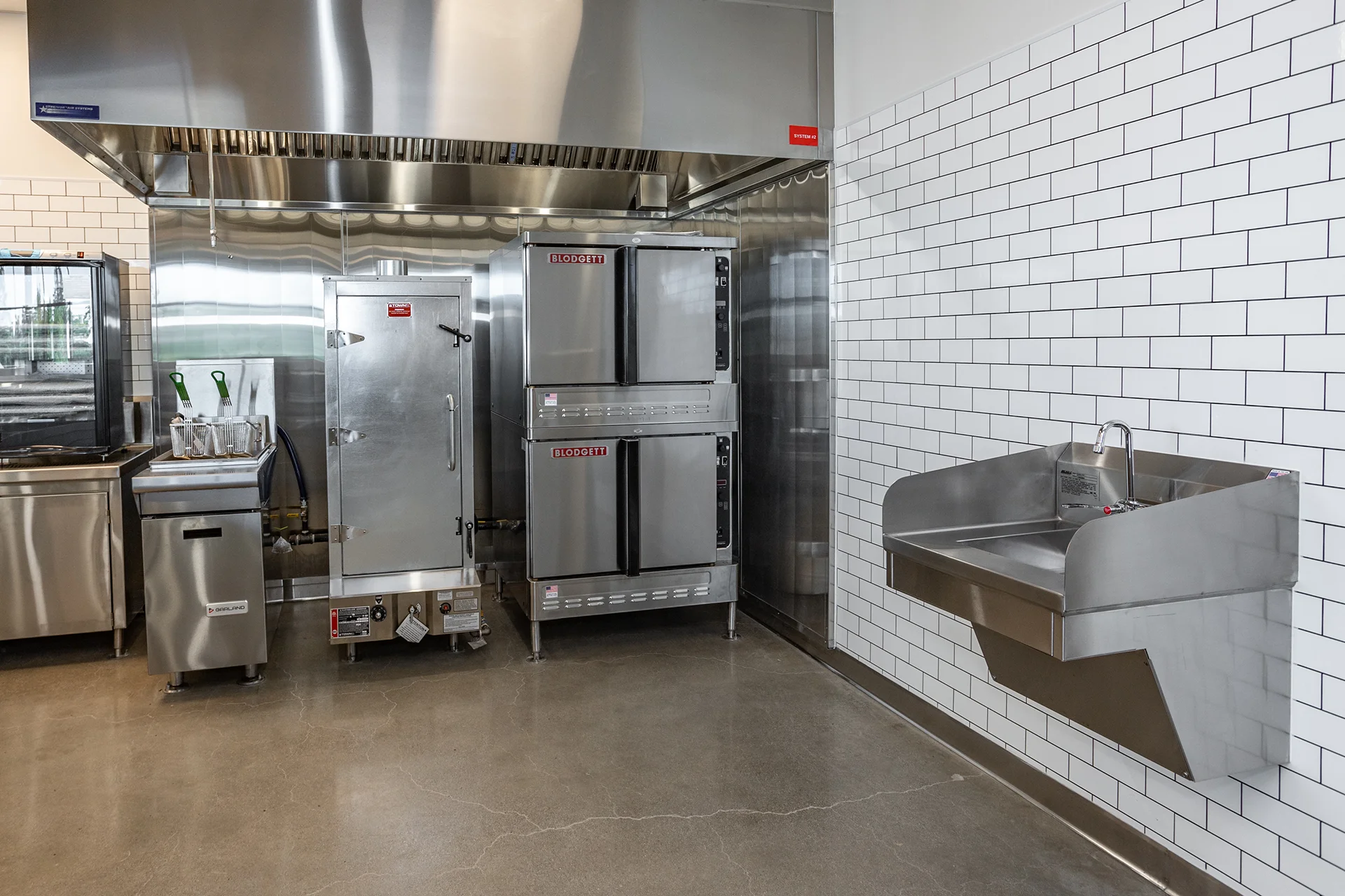 Heavy-duty baking and frying zone showcasing a double-stacked gas convection oven unit next to a standalone deep fryer with wire baskets, situated below an industrial exhaust canopy next to a wall-hung scrub sink.