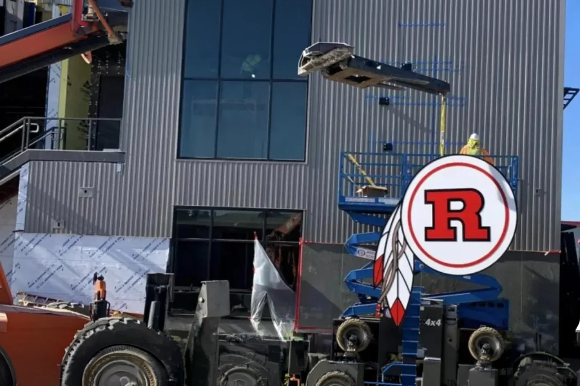Construction crew utilizing a commercial blue scissor lift to mount a circular graphic school emblem onto the corrugated metal panel siding of a multi-story institutional facility in Ripon, California, during the final build phase.