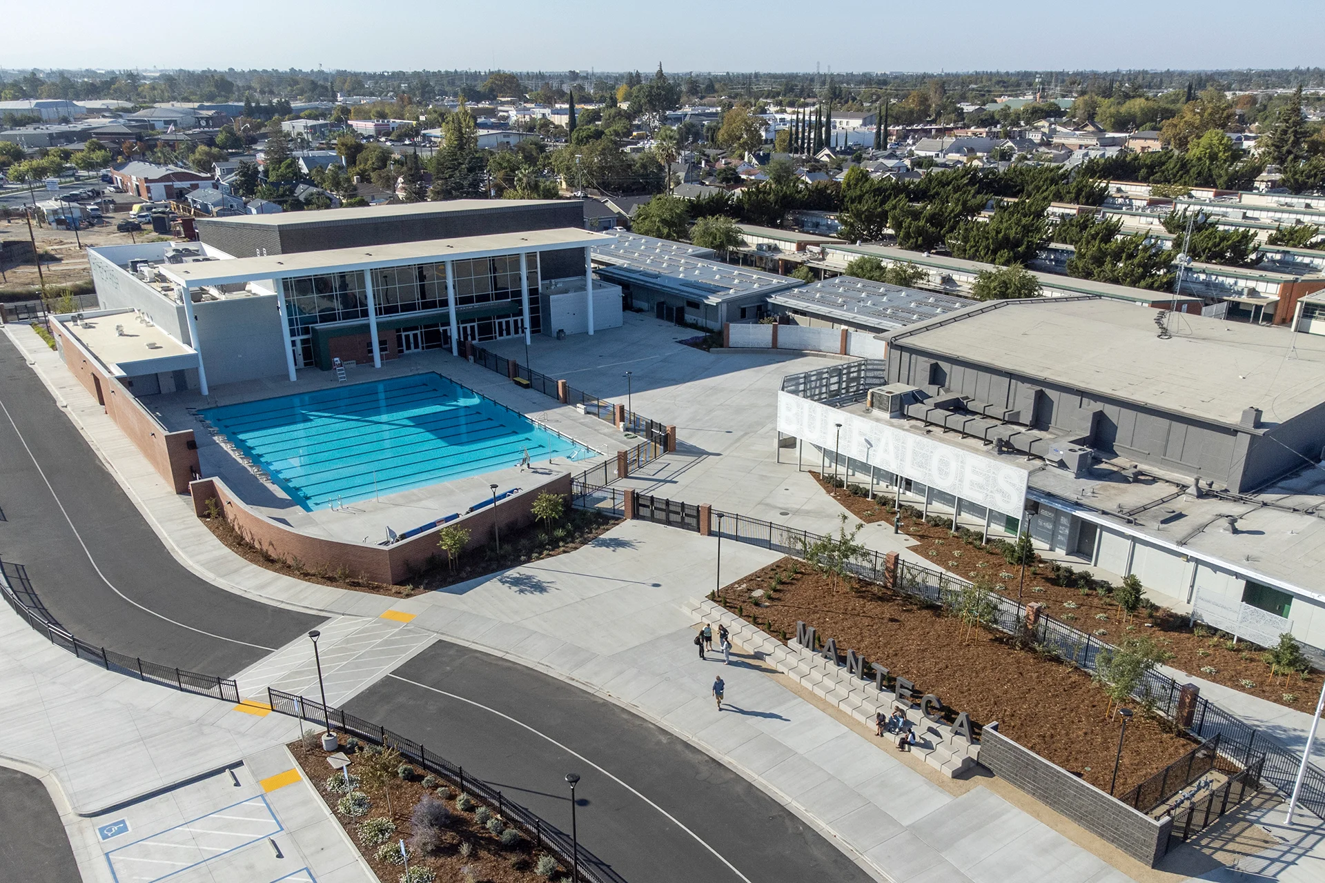 Comprehensive aerial view of an institutional sports plaza, showing an outdoor lap swimming pool flanked by a glass-fronted athletics pavilion, precast concrete monument steps serving as public benches, and an organized asphalt bus loop zone.