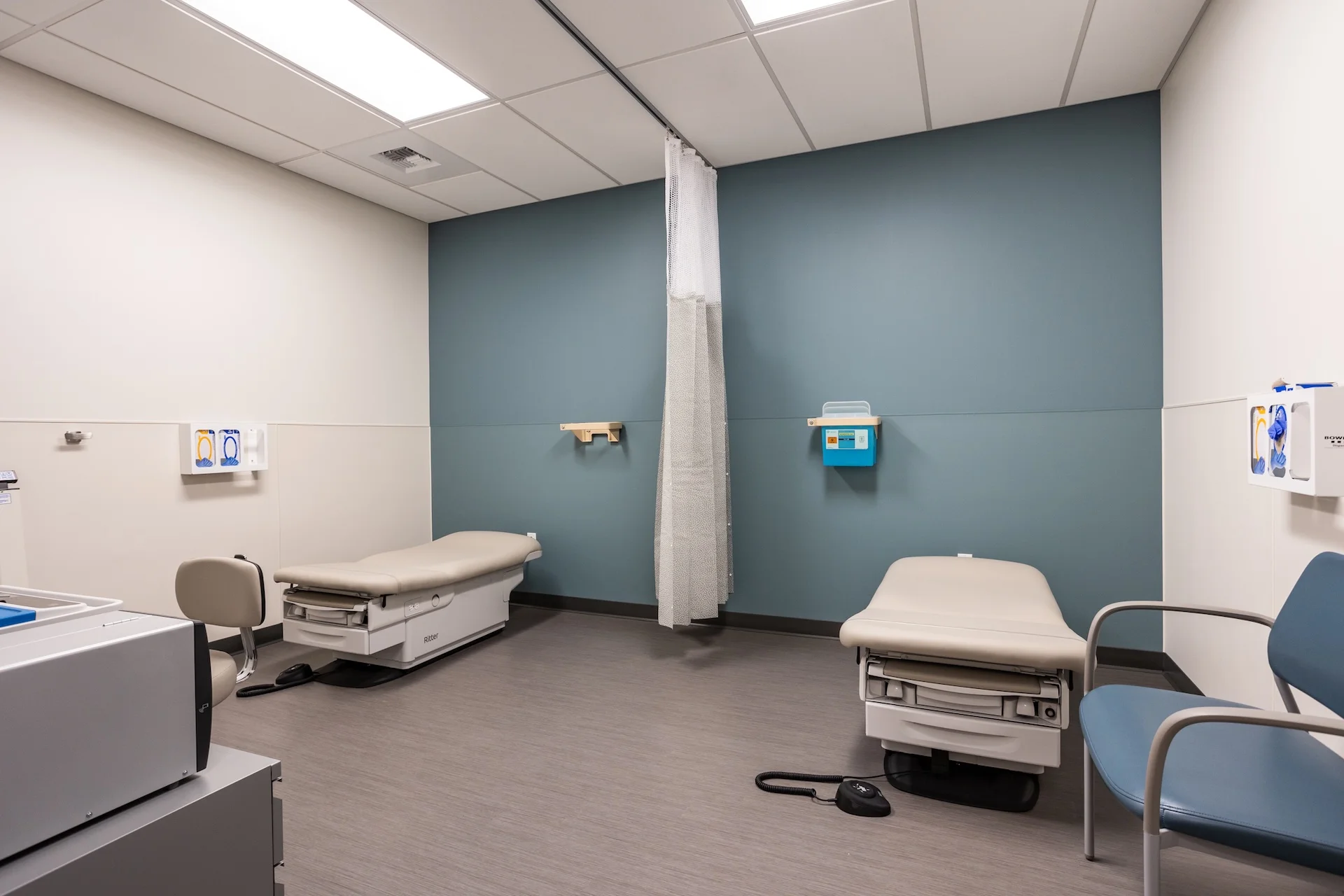 Double patient bay layout with two adjustable vinyl-upholstered medical examination beds separated by a central tracking privacy curtain, featuring a cool teal accent wall, wall-mounted glove dispensers, sharp disposal boxes, and vinyl plank floor.