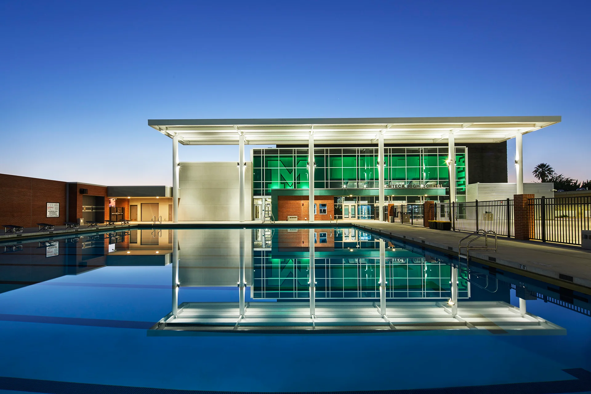 Evening exterior view of an institutional swimming pool facility in San Joaquin County, highlighting vibrant green interior lighting reflecting through a massive glass storefront wall into perfectly still blue water, framed by a lit metal shade canopy under a deep gradient twilight sky.