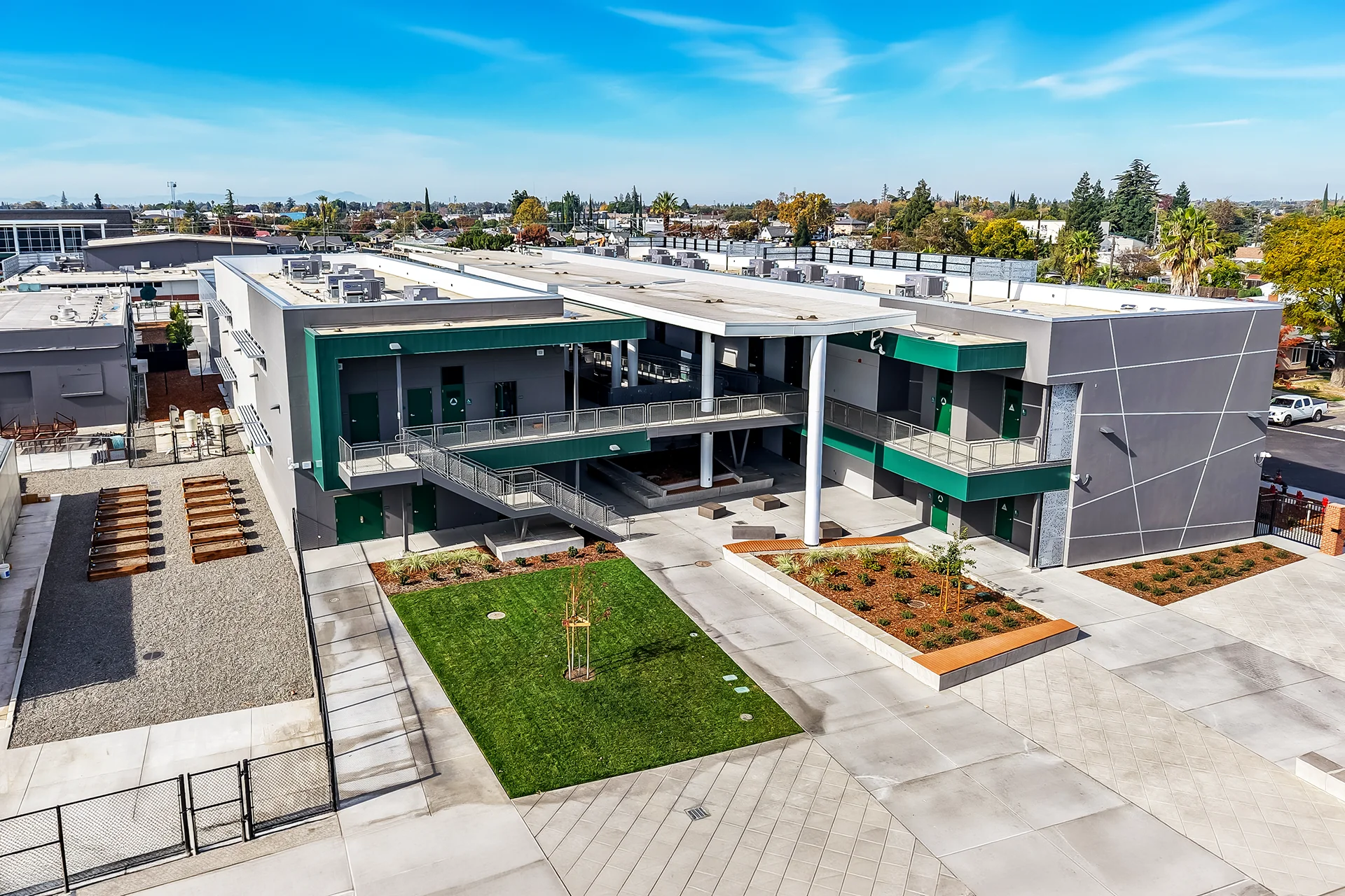 Architectural drone shot of a multi-level school quadrangle in San Joaquin County, highlighting grey and dark green stucco structural wings connected by open-air corridors with stainless steel woven wire mesh security railings, an exterior concrete staircase, and concrete plazas.