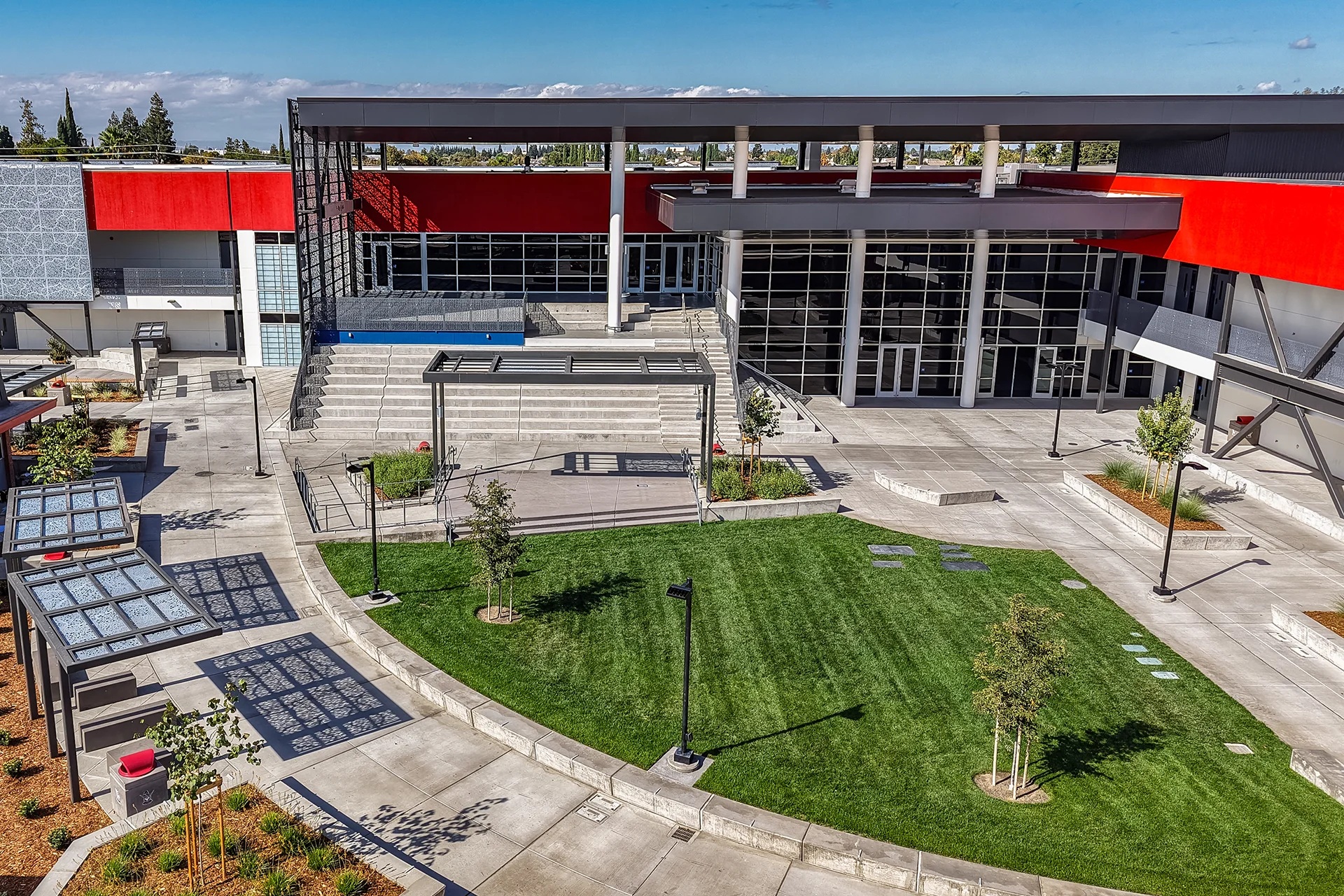 Architectural bird's-eye perspective of a school campus quad showing integrated concrete planter boxes, young landscaped trees, concrete flatwork pathways, a central grass turf lawn, and bold red building accents.