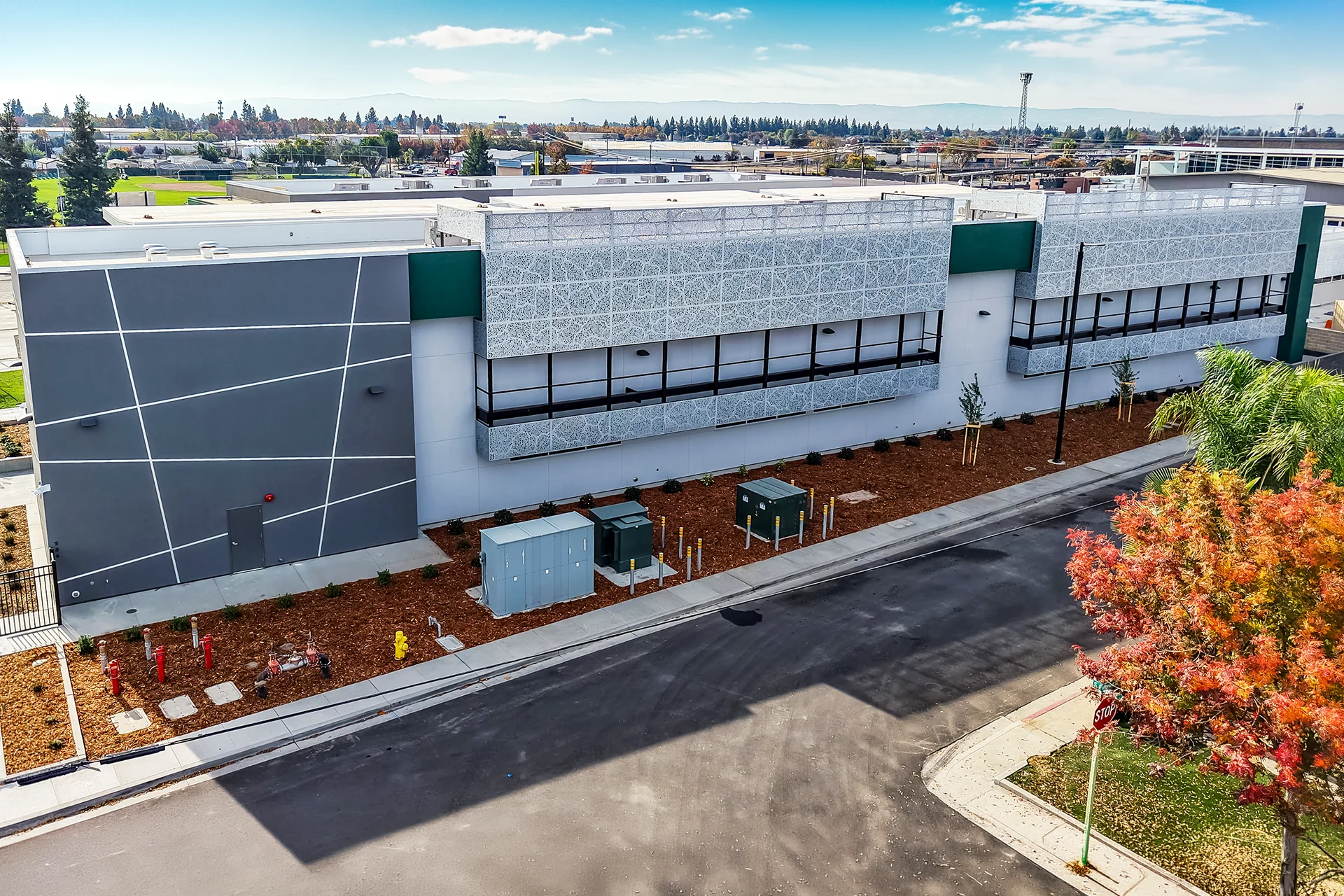 High-angle exterior drone view of a newly constructed modern academic building in Manteca, California, highlighting extensive laser-cut aluminum privacy screens over deep window channels, an accent wall with abstract white geometric lines, ground-level electrical transformers, and freshly laid wood bark mulch landscaping.