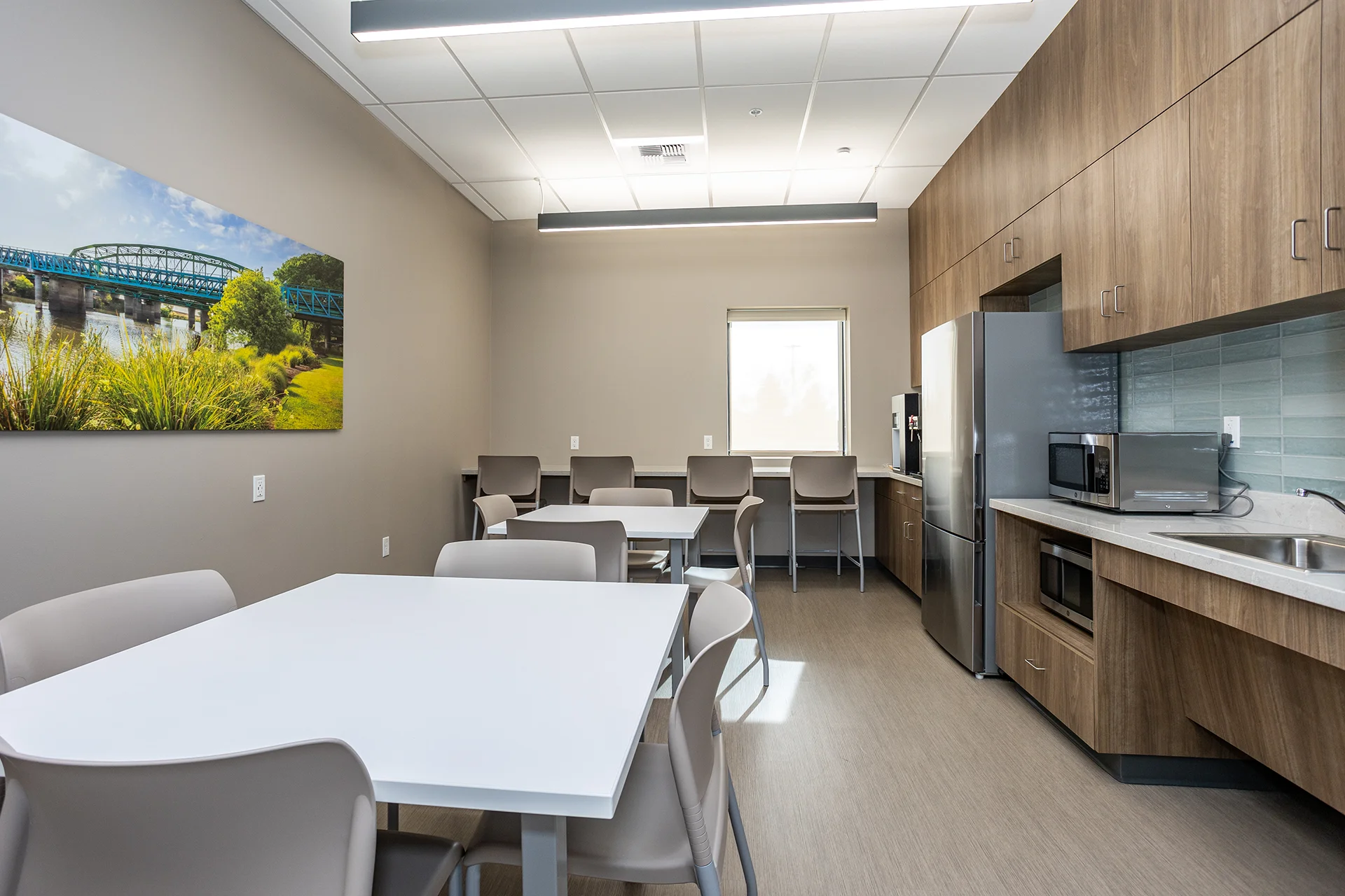 Clinical facility staff breakroom highlighting an integrated perimeter bar height counter with stools, a large wall-mounted landscape canvas print, premium walnut wood laminate cabinetry, white composite solid-surface countertops, and an energy-efficient stainless steel refrigerator.