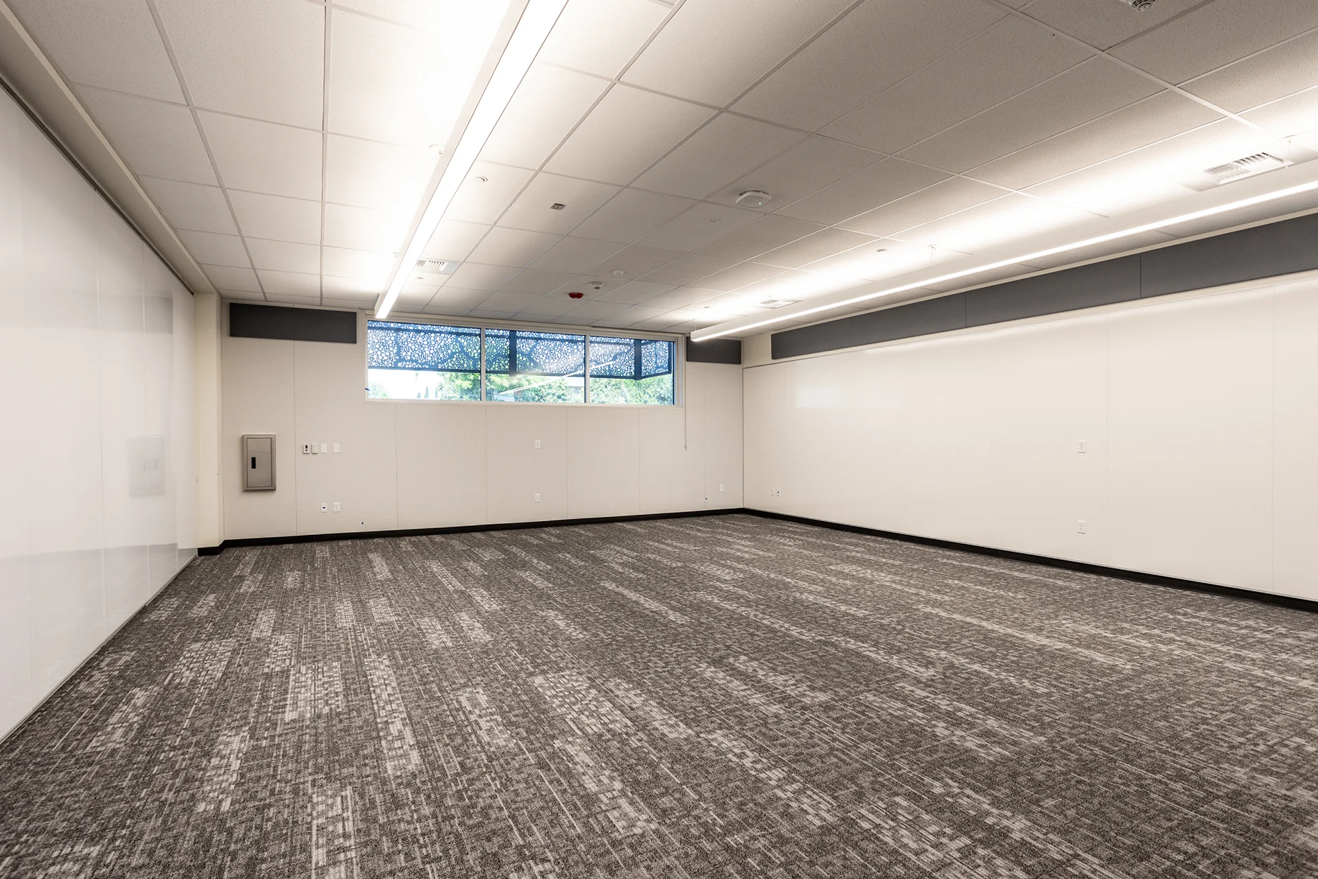 Freshly completed institutional classroom interior highlighting integrated white gloss presentation walls, multi-patterned carpet tiling, sleek surface-mounted linear LED lighting fixtures, and a glass privacy window.