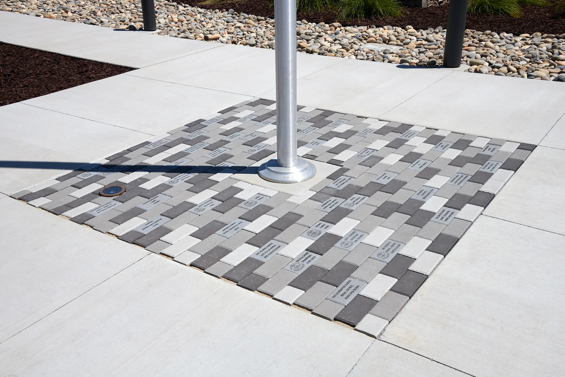 Close-up detail of the concrete walkway showing a decorative square grid pattern of multi-colored grey and white brick pavers engraved with community names surrounding a structural brushed aluminum flagpole base.
