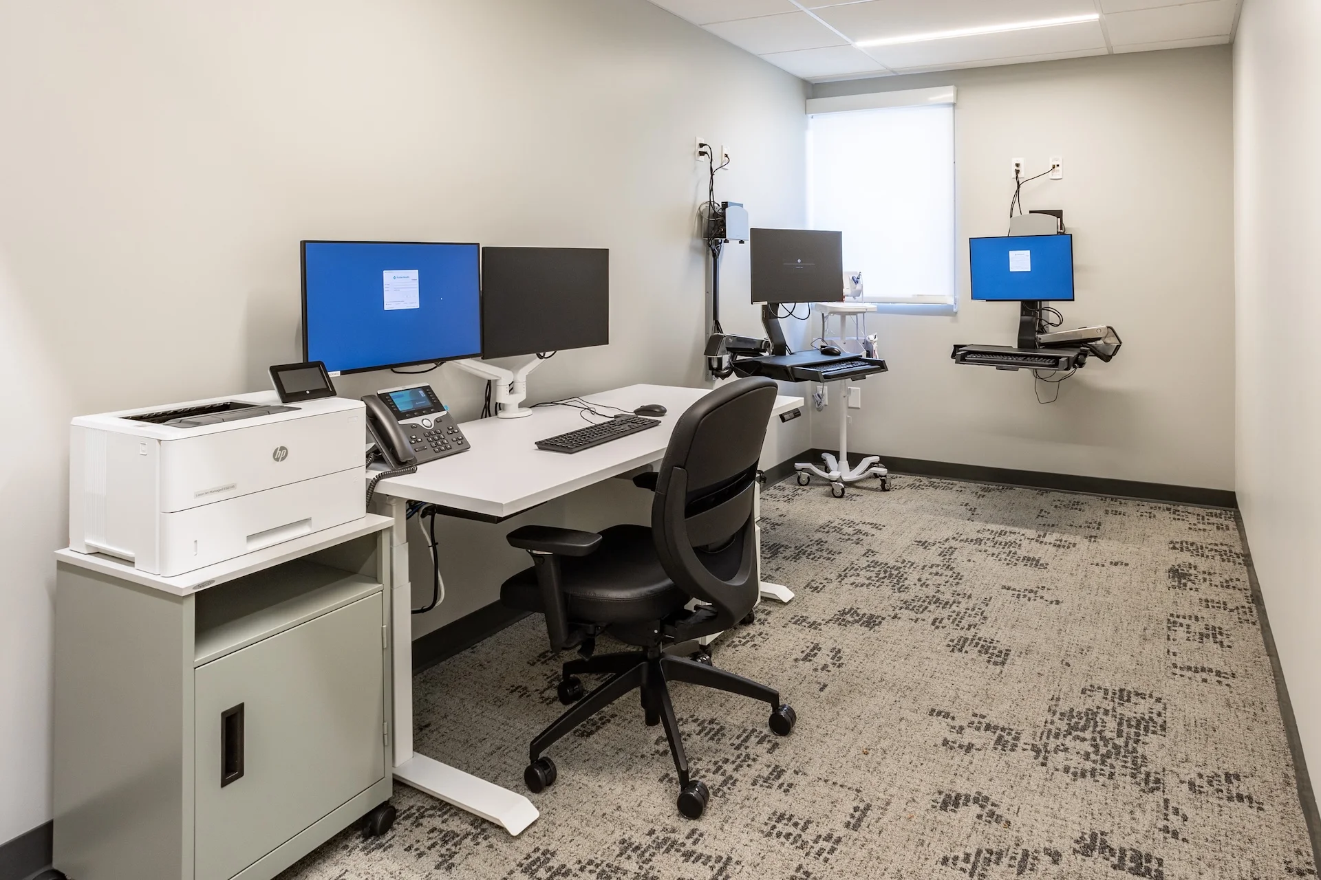Modern medical administrative alcove containing a motorized white sit-to-stand desk, dual monitor mounts on articulating arms, an ergonomic rolling task chair, a network printer station, and sound-dampening commercial carpet tile with an organic geometric weave.