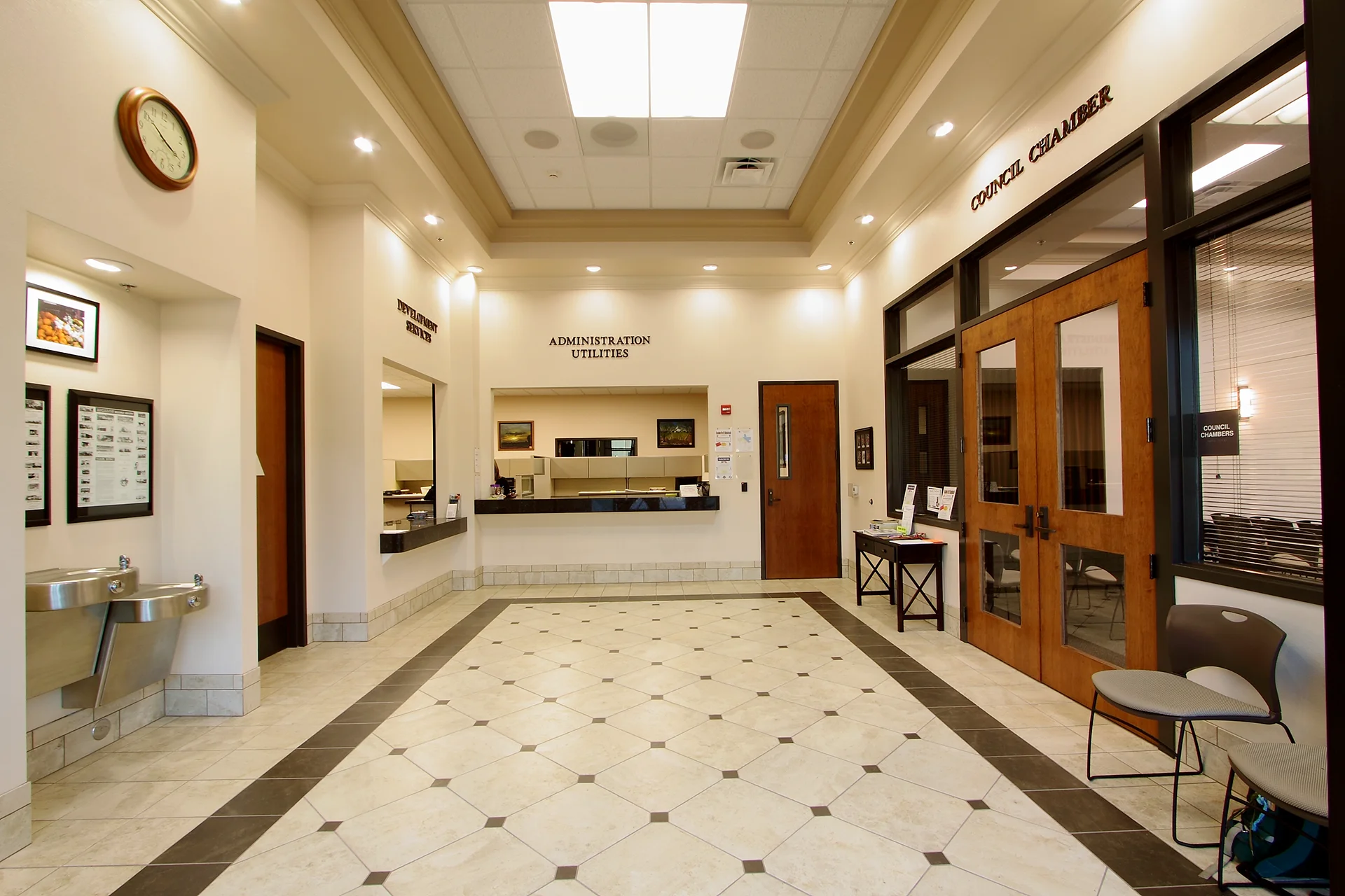 Main public lobby of Escalon City Hall highlighting the polished ceramic tile flooring with decorative borders, built-in service windows for Administration and Utilities, a stainless steel water fountain, and double wood-frame glass doors leading into the council chambers.