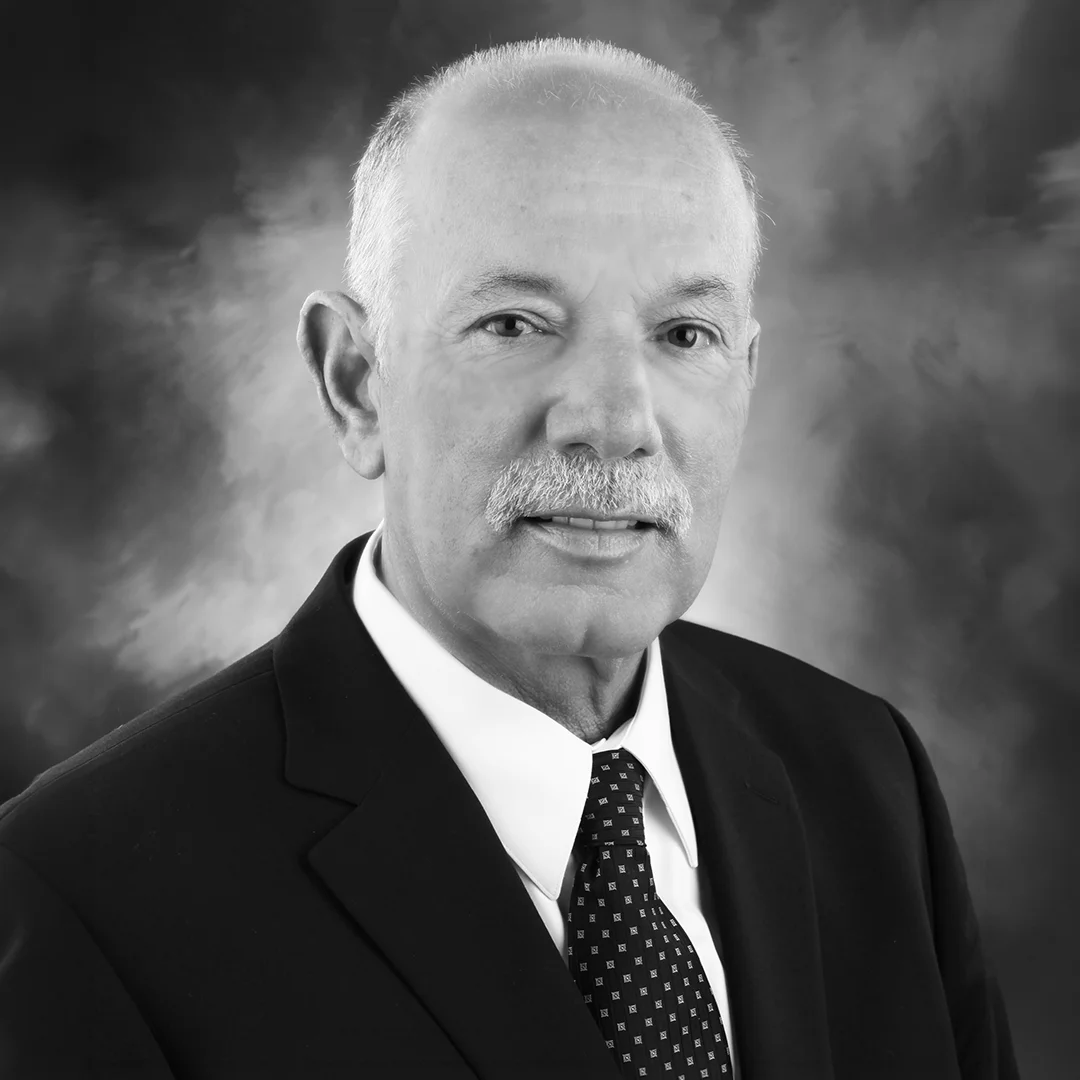 Professional black-and-white corporate headshot of a smiling senior executive with a mustache, dressed in a dark business suit, white dress shirt, and patterned tie against a textured studio backdrop.