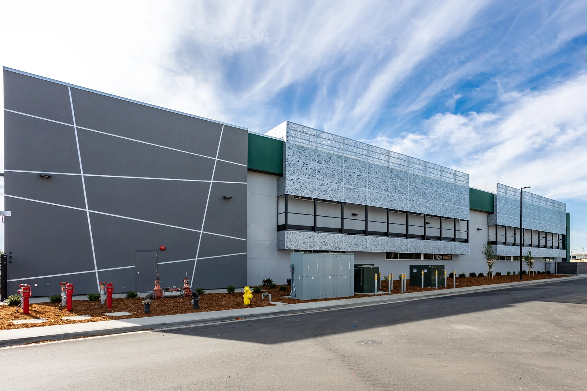 Profile view of a modern school campus building exterior in San Joaquin County, showcasing a gray stucco accent wall with abstract white reveal lines, ground-level utility infrastructure with commercial backflow preventers, green and white paneling, and architectural metal canopy screens under a bright blue sky.
