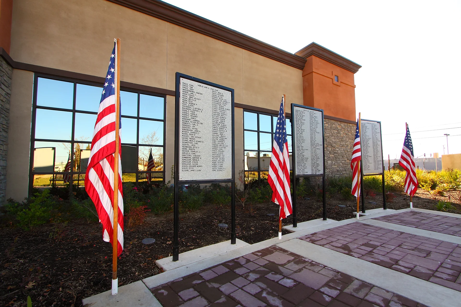 Outdoor veteran's memorial display at the Waterford municipal complex, showcasing three black metal panels listing names of military service members, bordered by concrete pavers and flanked by small wood poles flying American flags.