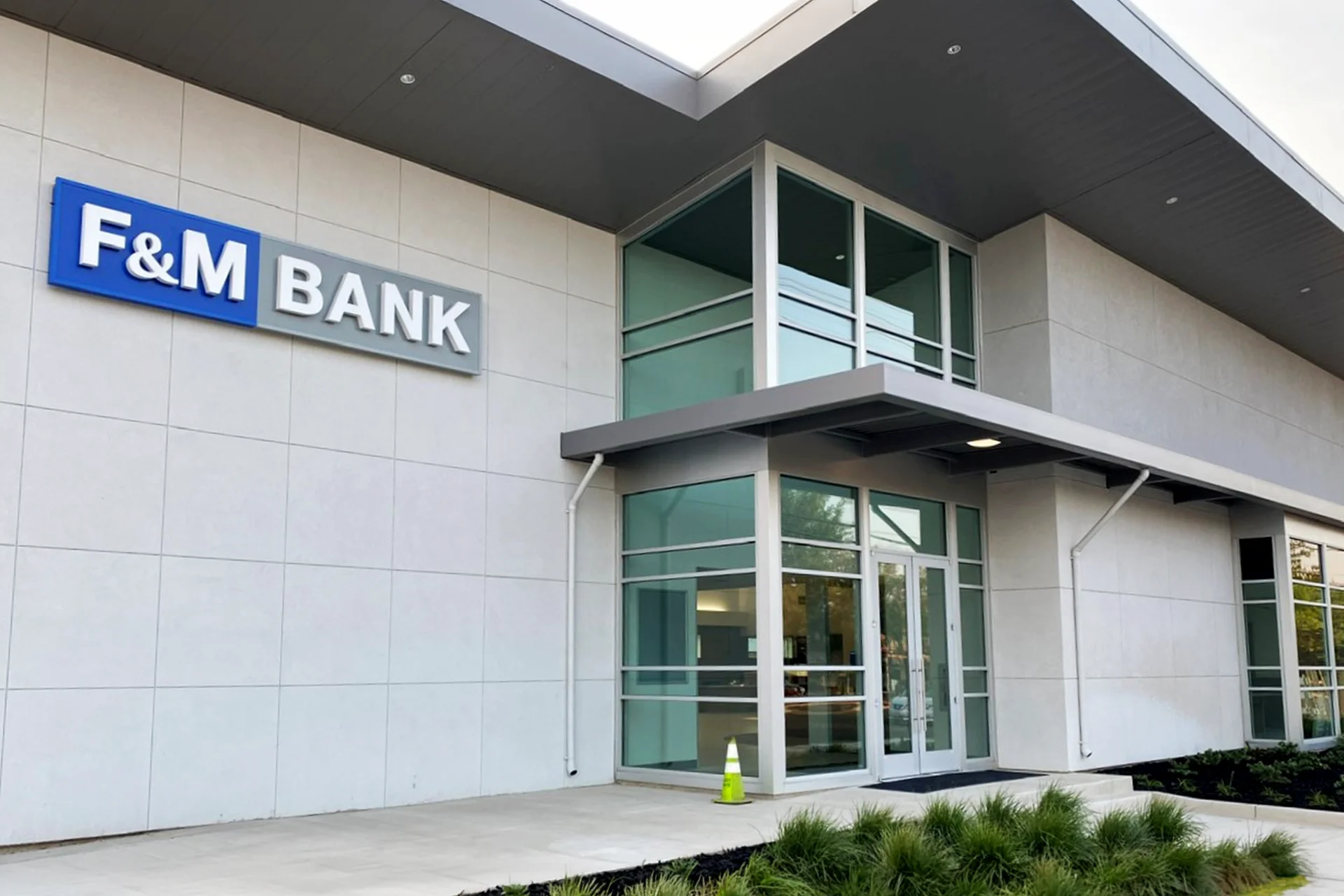 Exterior front entryway detail of the F&M Bank branch in Turlock, California, showcasing large square ceramic wall tiles, an illuminated blue brand sign, a two-story glass storefront vestibule, and a deep-set architectural metal overhang.