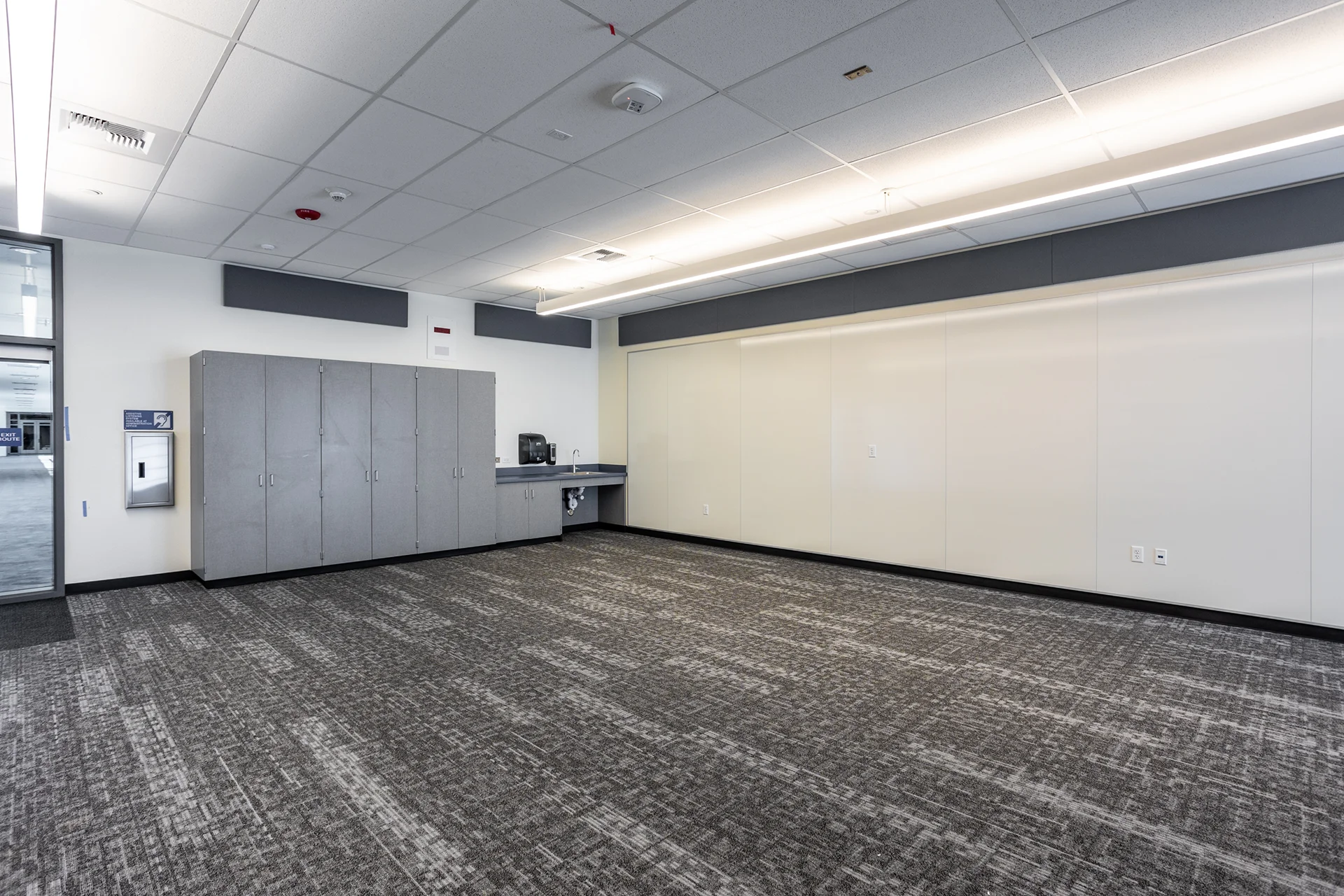 Modern commercial school room showing low-profile linear LED drop lighting, custom gray laminate storage millwork, an integrated sink station, patterned gray commercial carpeting, and premium wall-to-wall white presentation panels.