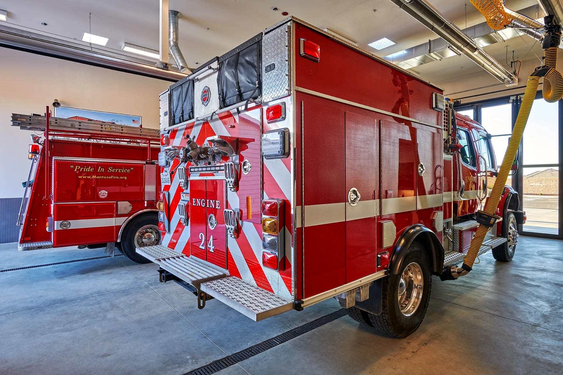 Interior perspective of the station apparatus bay showing the rear profile of fire Engine 24 parked on a sealed concrete floor with active yellow overhead exhaust extraction hoses connected to the vehicle.
