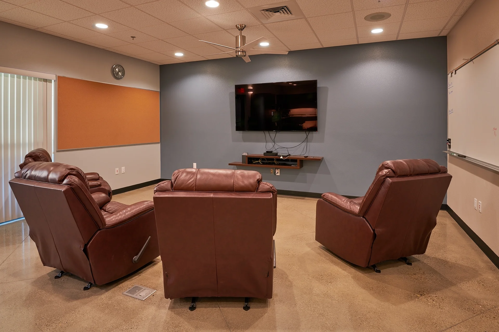 Comfortable dayroom area for fire station personnel featuring brown leather reclining chairs, a wall-mounted flat-screen television, a large corkboard, a dry-erase whiteboard, and stained concrete flooring.