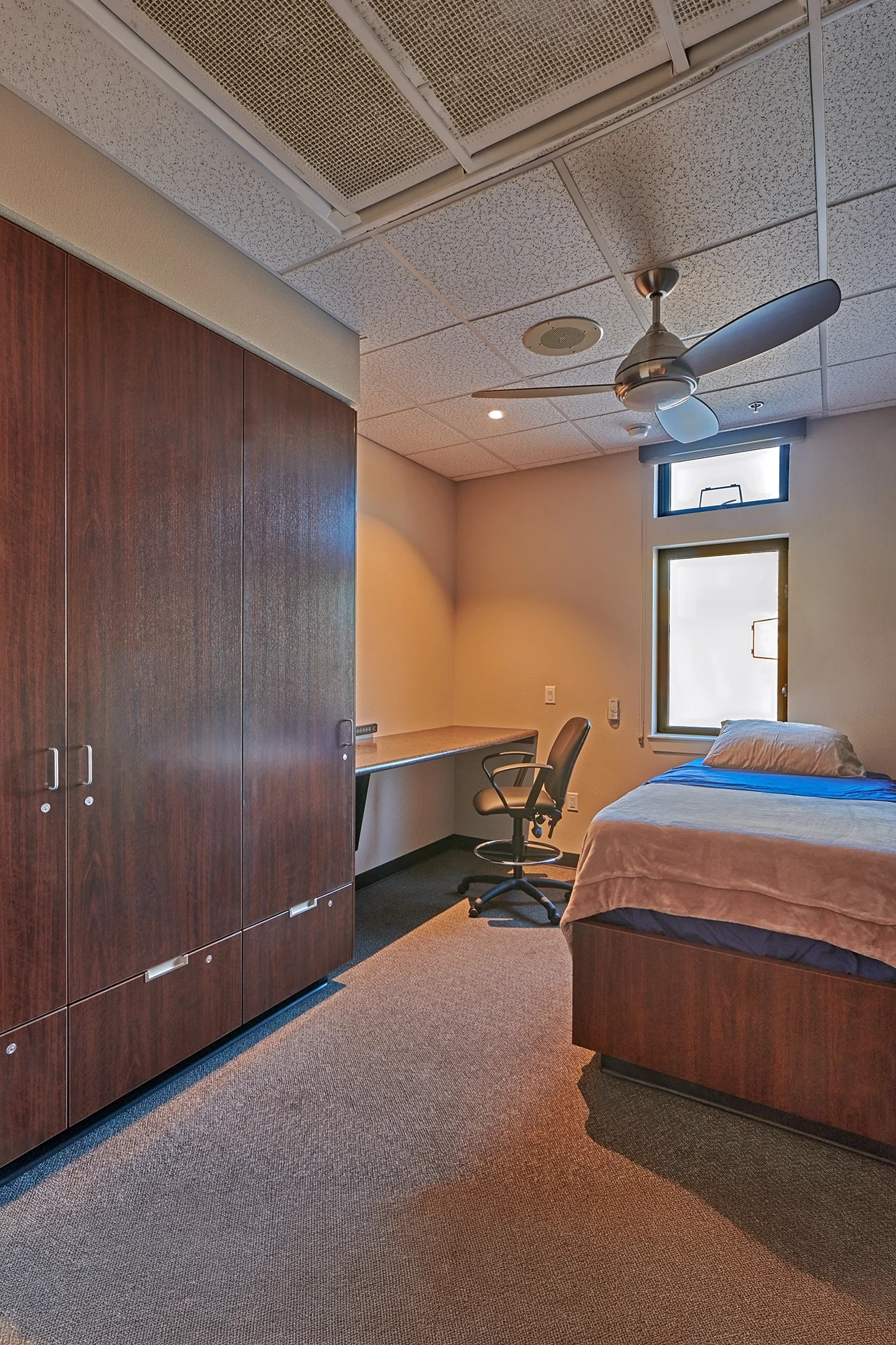 Private crew sleeping quarters inside the Manteca station showcasing a single bed with blue bedding, a built-in laminate desk system with an ergonomic office chair, a large wooden wardrobe unit, a ceiling fan, and high privacy windows.