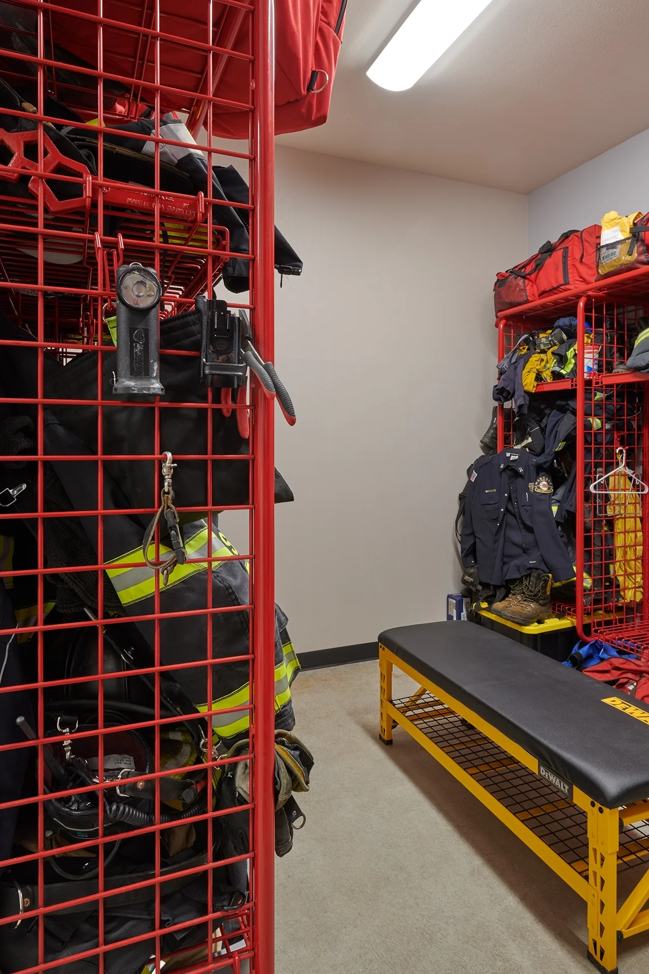 Specialized station storage room featuring heavy-duty red wire mesh lockers holding protective first responder turnout gear, coats, helmets, and duffel bags alongside a yellow-framed DeWalt utility bench.