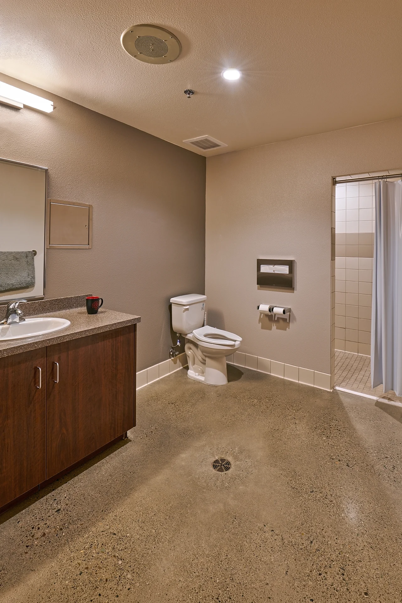 Commercial-grade station bathroom installation featuring a dark wood vanity with a solid surface countertop, a standard white ceramic toilet, a central floor drainage grate in the sealed concrete floor, and an adjacent walk-in shower stall with white tiling and a blue curtain.