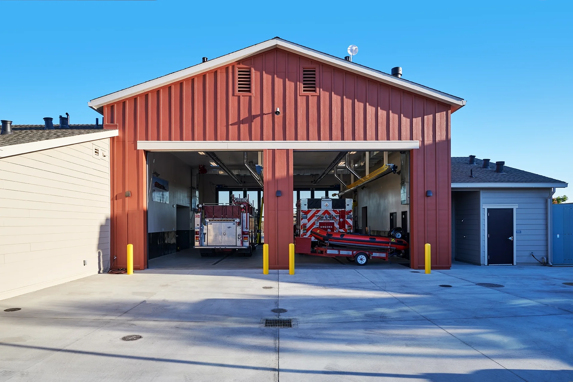 Symmetrical view looking straight into the open garage bays of the station house, showing an emergency rescue boat trailer and fire engine parked on the reinforced concrete slab floor under a clear blue sky.