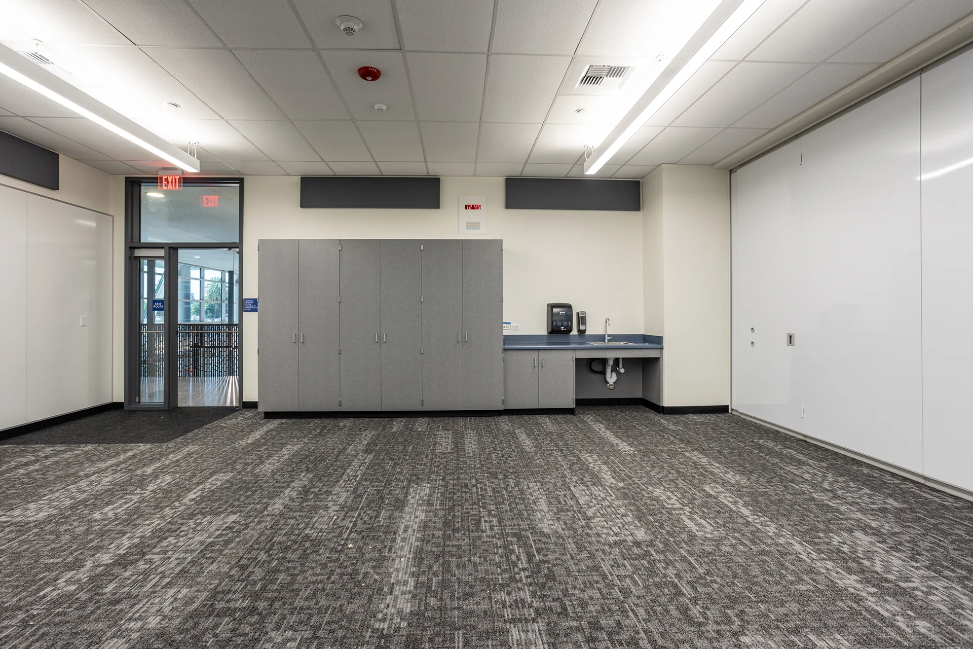 Campus educational wing interior featuring heavy-duty gray patterned commercial carpeting, custom gray laminate millwork wardrobes, a perimeter hand-washing station, and a wide white sliding divider partition wall.