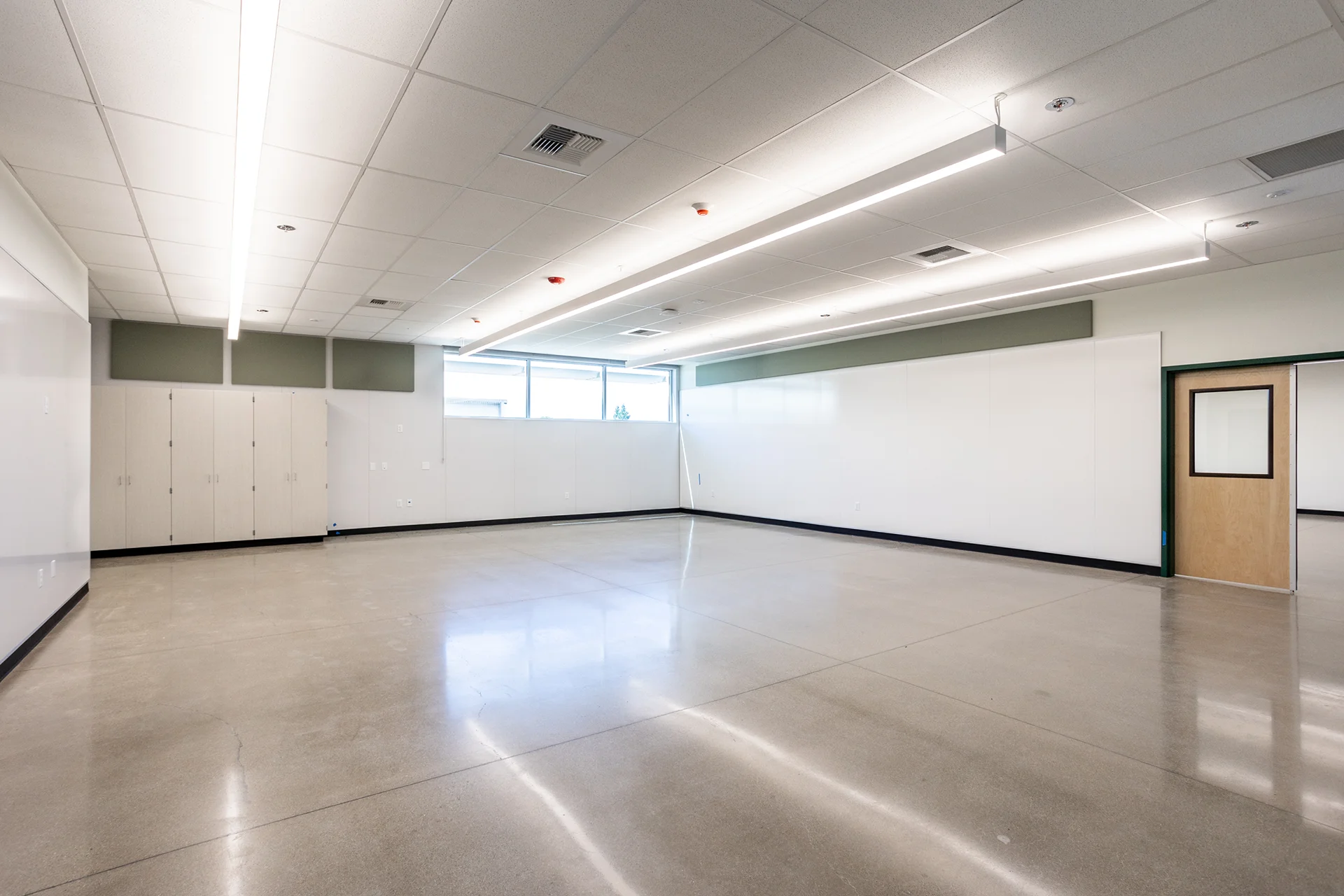 Luminous empty classroom interior in San Joaquin County, highlighting premium polished concrete floors reflecting linear LED flush light fixtures, modular white wall panels, integrated sound dampening baffles, and a maple veneer door with view panel.