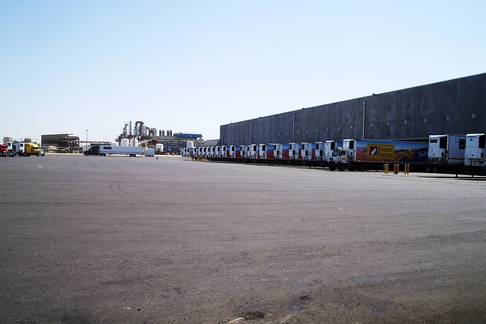 Truck loading area at the Foster Farms National Chicken Distribution Center with refrigerated trailers parked at warehouse loading docks and industrial processing facilities in the background.