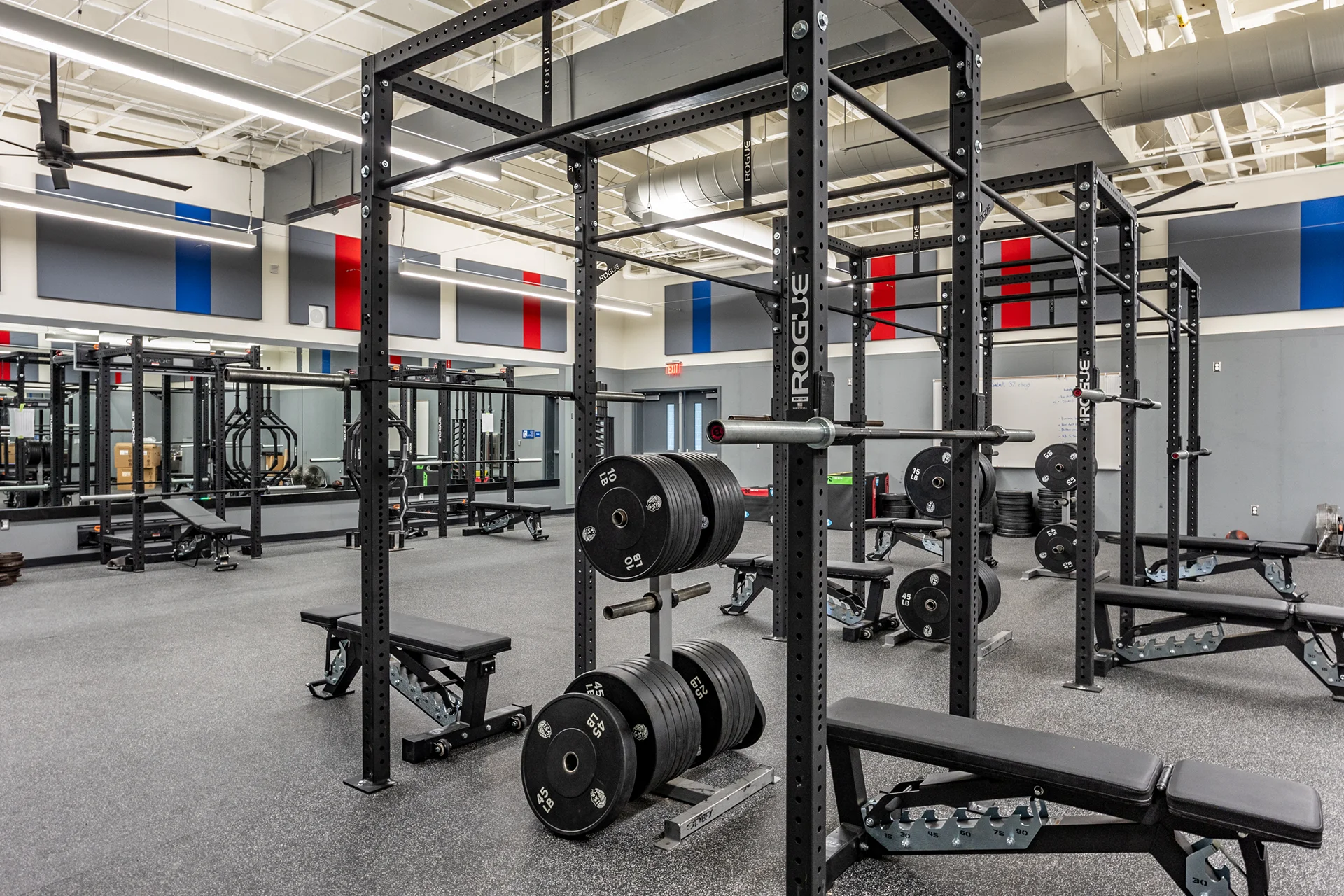High school fieldhouse weight training center highlighting multiple modular steel squat racks, Olympic barbells with bumper plates, adjustable workout benches, a white dry-erase board, and acoustic wall panels.