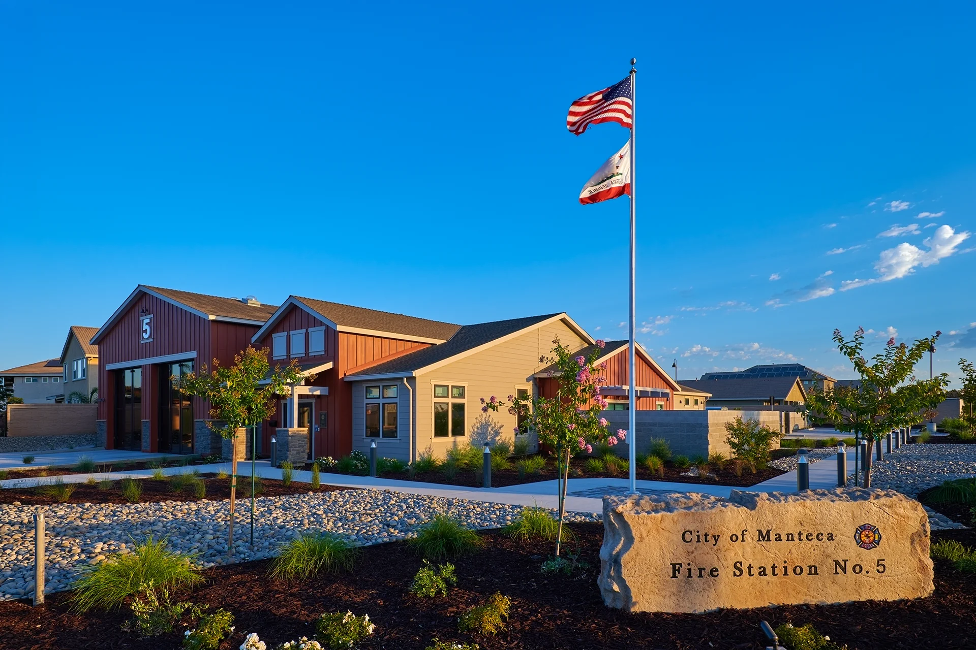 Sunny daytime view of the completed City of Manteca Fire Station No. 5 building, showcasing red and tan horizontal siding, an engraved natural stone monument sign, a flagpole flying American and California flags, and stone river rock landscaping.