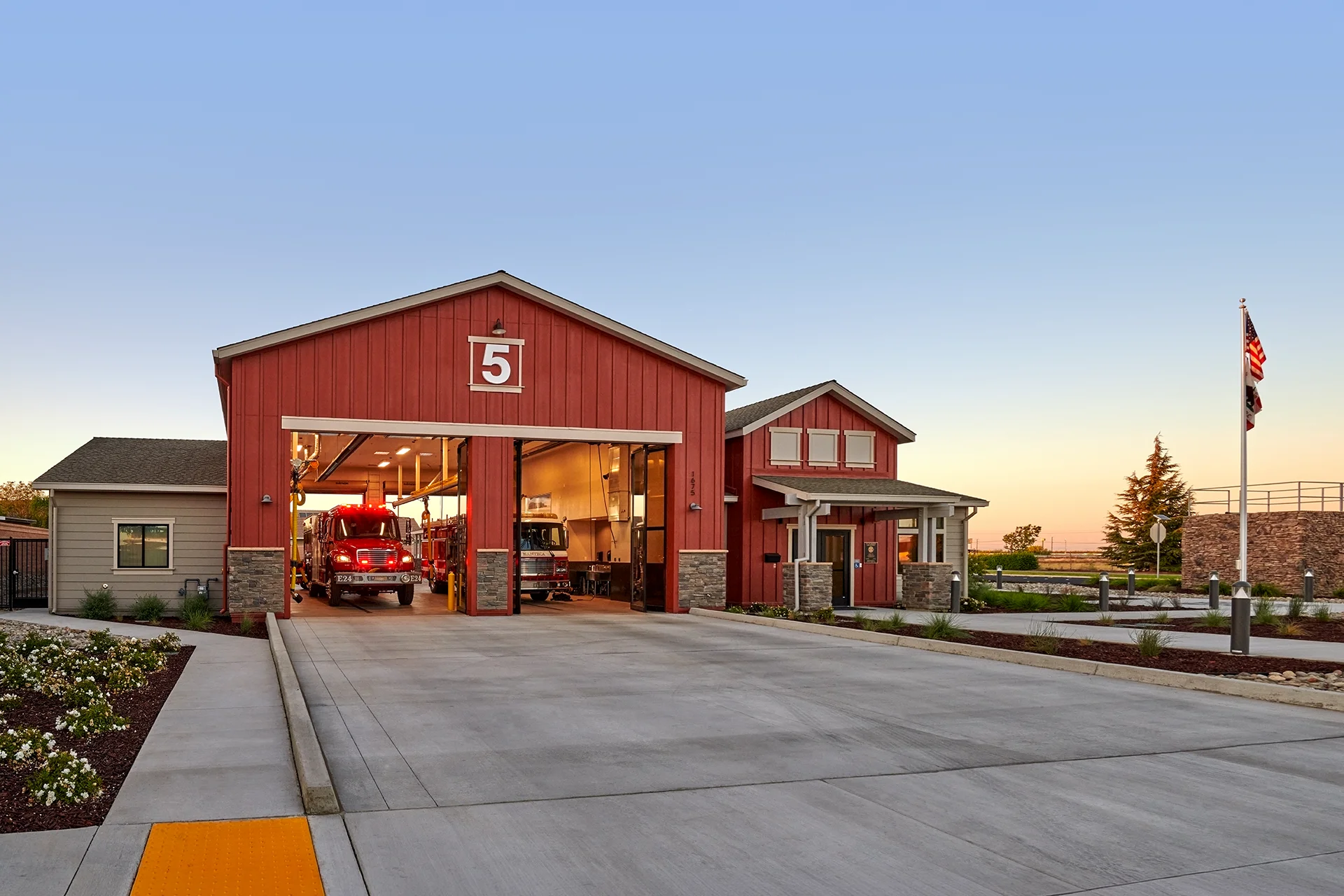 Dusk architectural photography of the station house in Manteca, California, highlighting the illuminated glass garage bay doors, parked response vehicles, manufactured stone pillar bases, and an American flagpole.