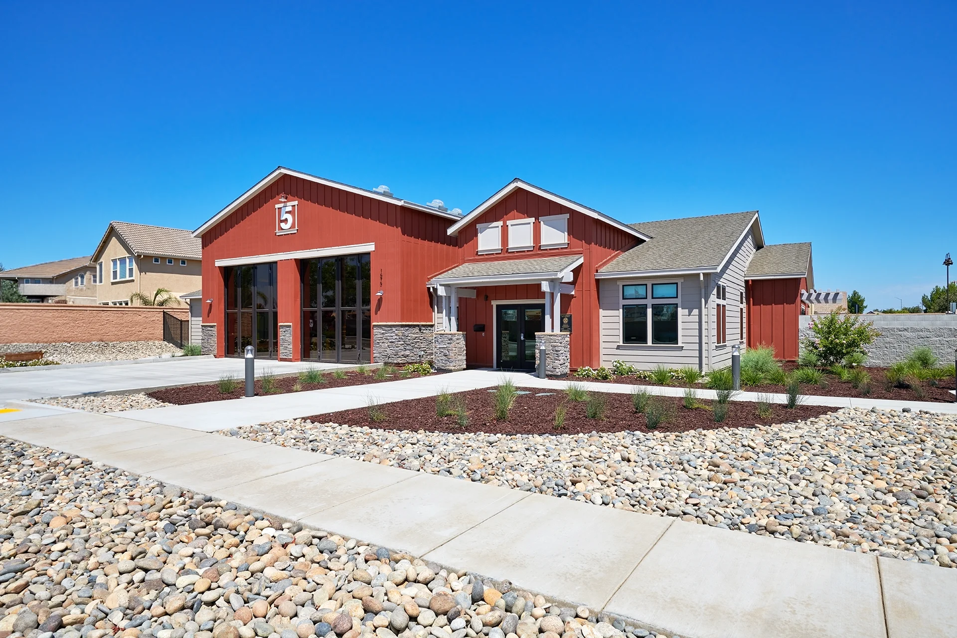 Wide-angle daytime shot of the completed civic facility, highlighting the red board and batten siding of the apparatus bays, a covered main entryway with stone veneer pillars, and a river rock landscaping bed bordering the concrete sidewalk.