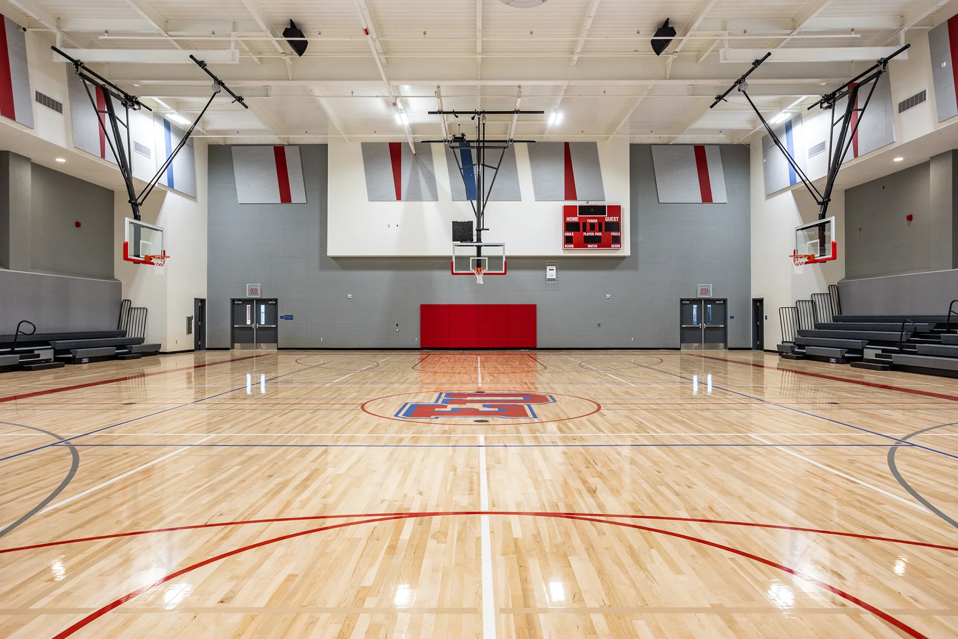 Comprehensive indoor basketball court renovation in Manteca, California showing high-gloss maple wood floors, retracted wall bleacher seating, gray sound-absorbing brick walls, red padding, and a digital wall scoreboard.