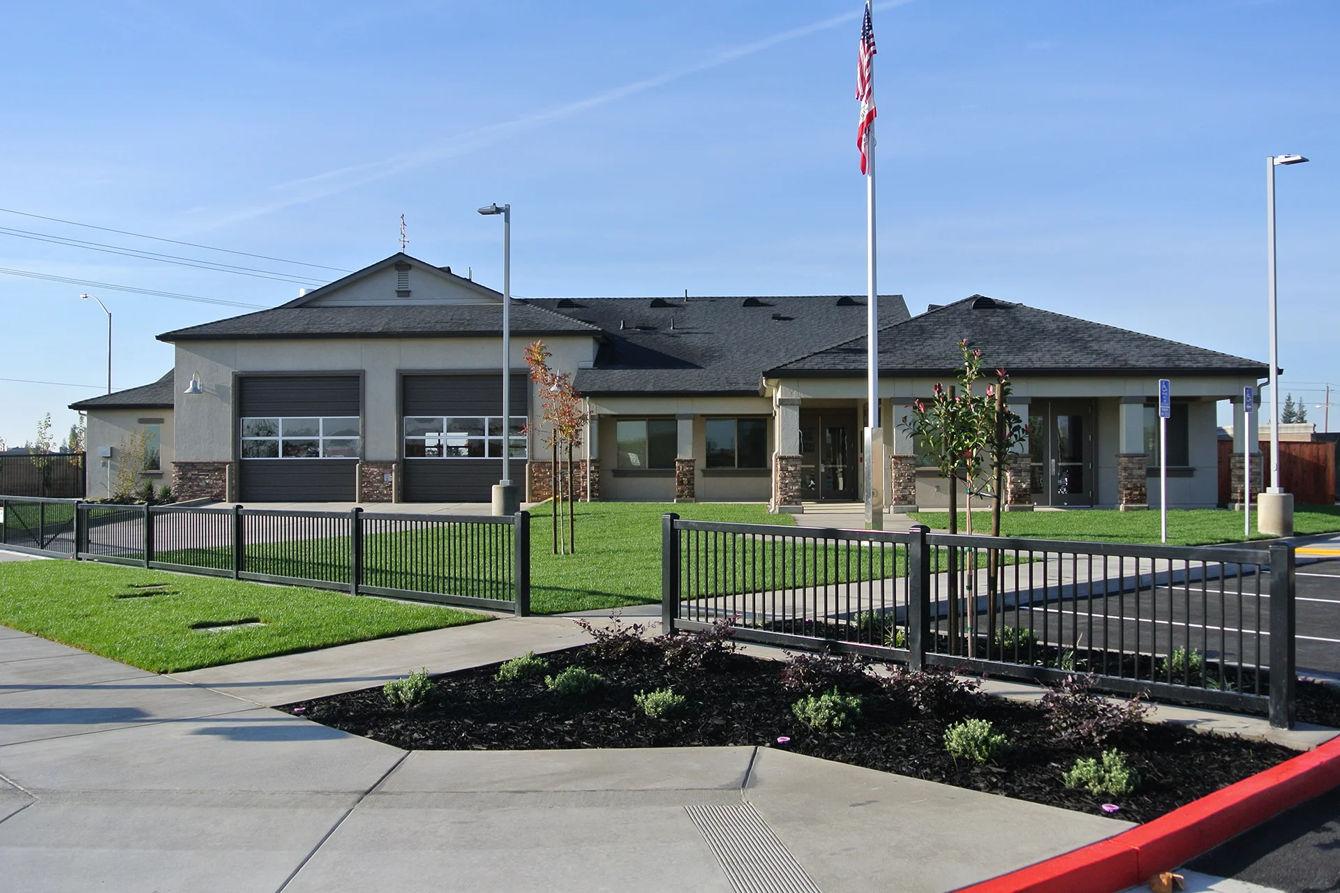 Wide daytime shot of the completed fire station construction under a clear blue sky, showing a landscaped green lawn behind a black metal perimeter security fence, a tall aluminum flagpole displaying the American and California flags, and a concrete driveway.
