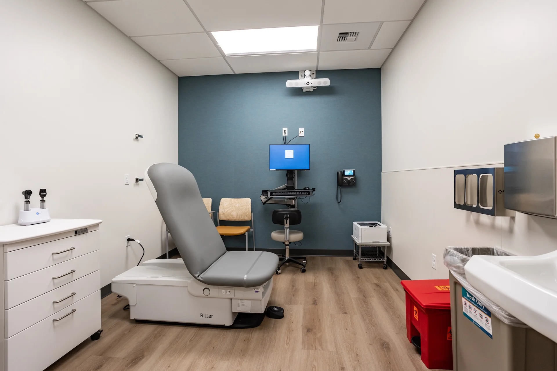 Specialized clinical exam space showing an adjustable gray vinyl examination bed, a rolling mobile supply cart with steel drawers, a hands-free handwashing station, wood-look luxury vinyl tile flooring, and a slate-blue wall backdrop featuring an integrated digital healthcare interface terminal.