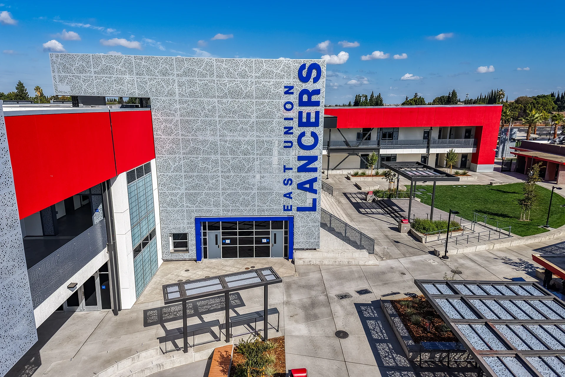 Architectural drone photo of an educational plaza highlighting a central entrance tower with custom blue lettering, geometric metal shade pergolas, landscaped planters, and a manicured lawn quad.