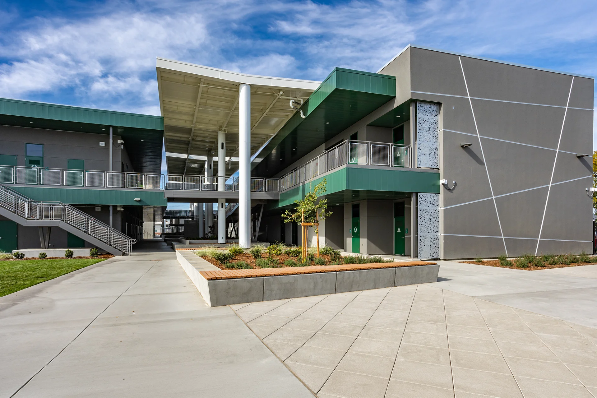 Up-close view from a concrete courtyard looking towards a massive covered open-air steel canopy structure spanning between two-story modern school buildings in Manteca, California, featuring laser-cut metal privacy screens and wood-topped concrete seating benches.