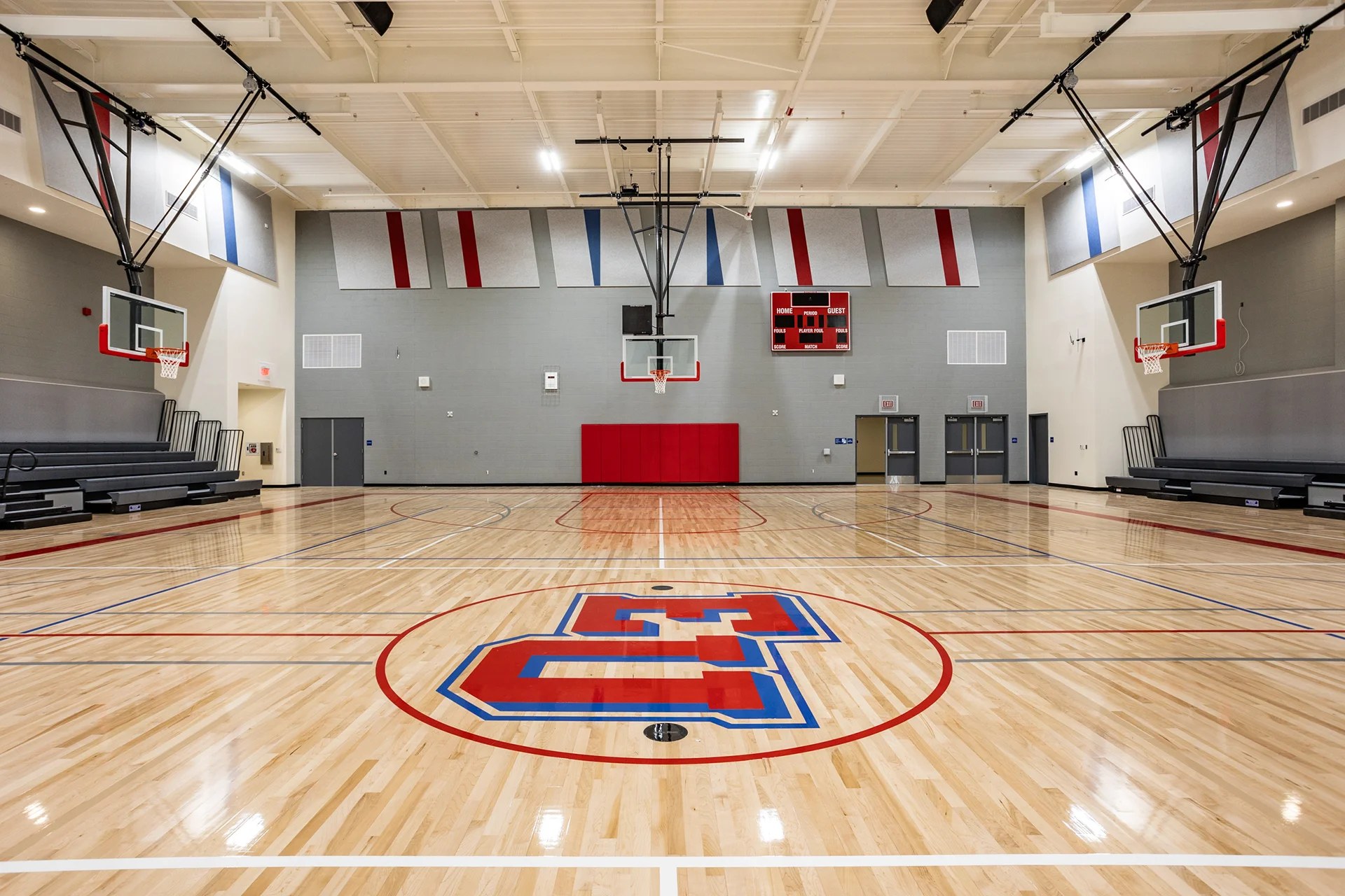 Symmetrical view of a modern school gym renovation highlighting a high-gloss maple wood floor with a large painted center court emblem, a digital scoreboard, structural steel ceiling trusses, and gray brick walls.