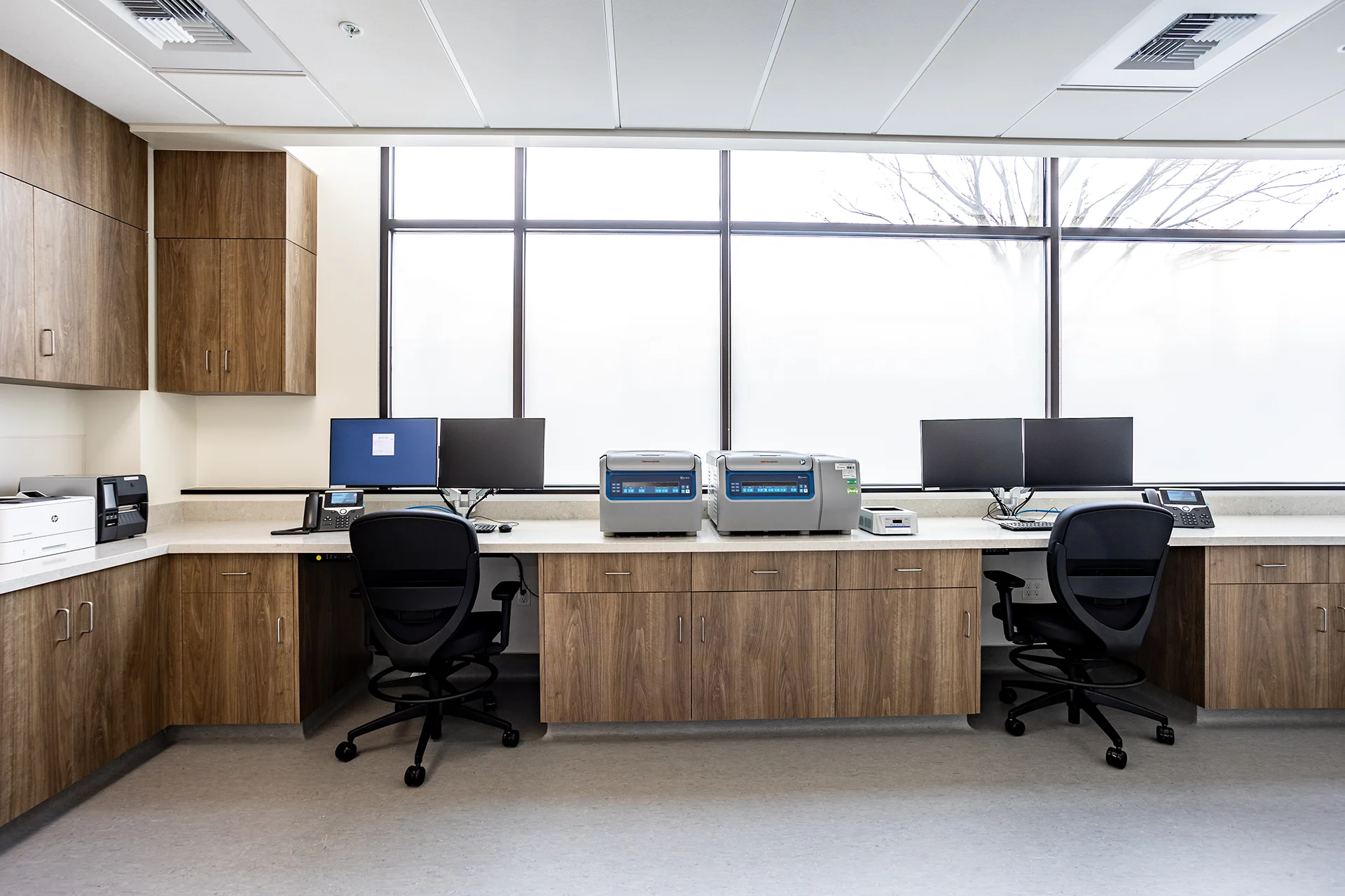 Main clinical laboratory processing counter featuring speckled white composite solid-surface tops, custom walnut laminate millwork cabinets, dual ergonomic lab stools, desktop computer workstations, and professional benchtop centrifuges.